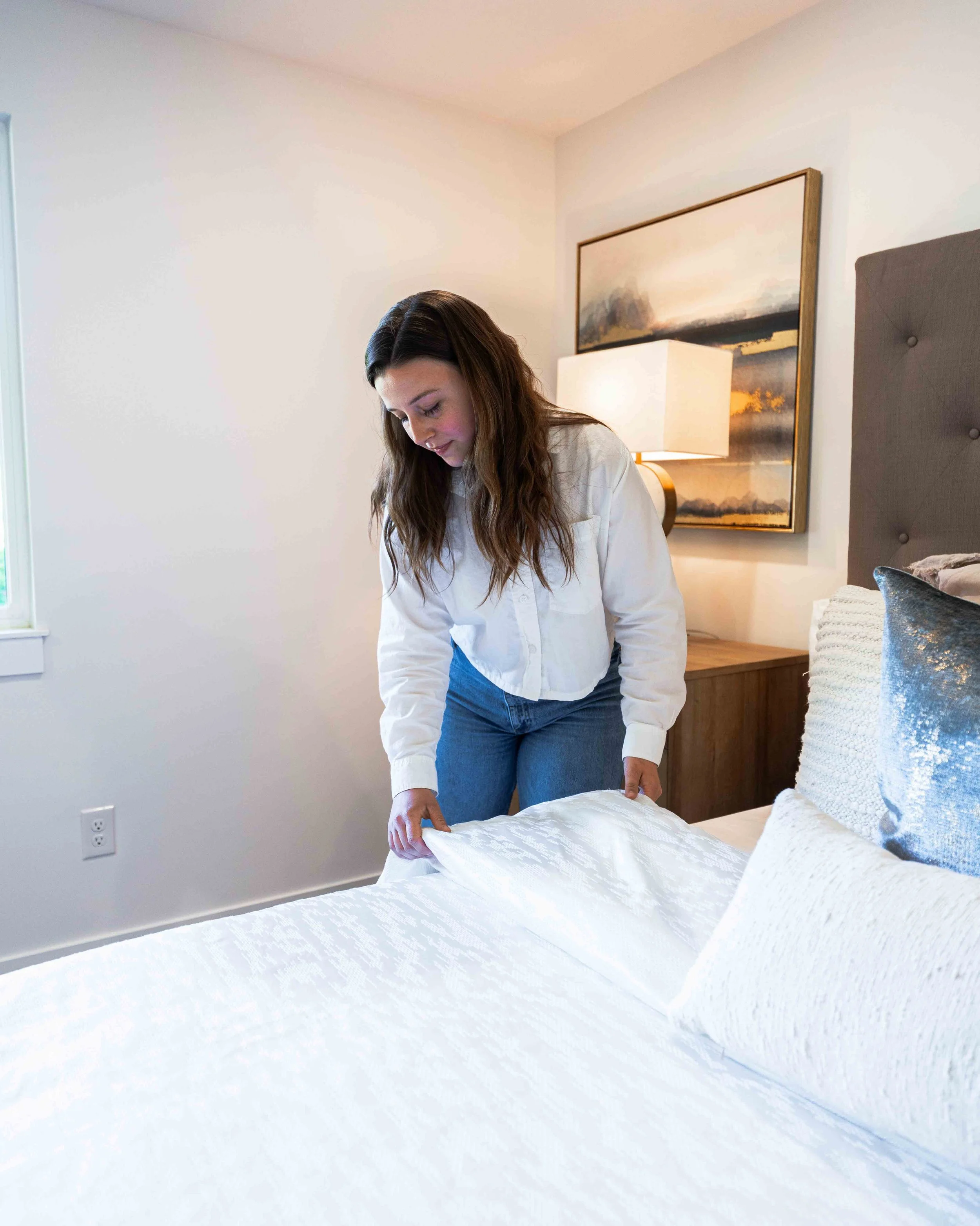 Woman making a bed with white quilt in a bedroom with gray headboard, painting, and lamp.