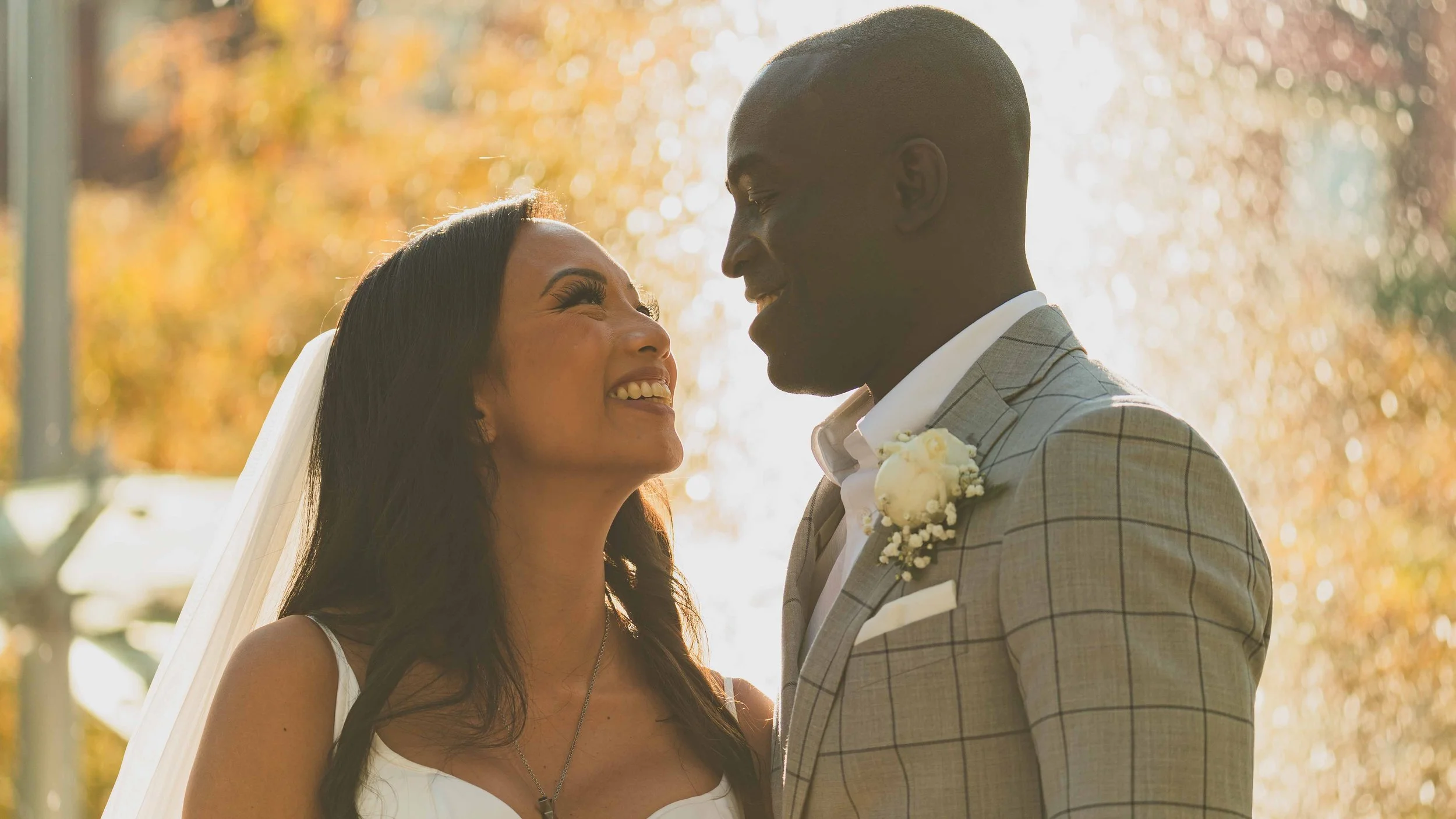 A bride and groom smiling and looking at each other outdoors during sunset with fall trees in the background.