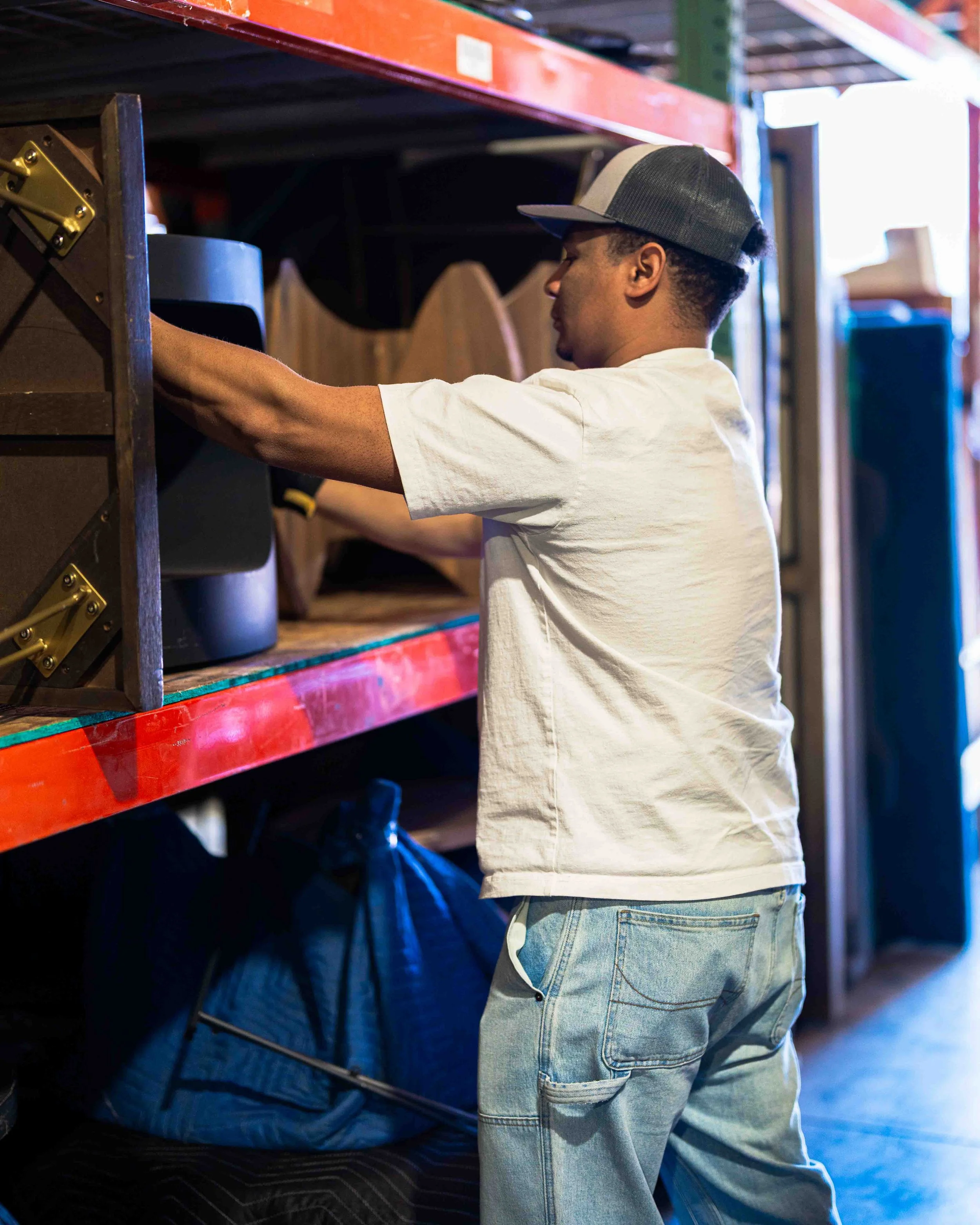 A young man with a cap and casual clothes is looking at and reaching towards storage shelves in a hardware or warehouse store.