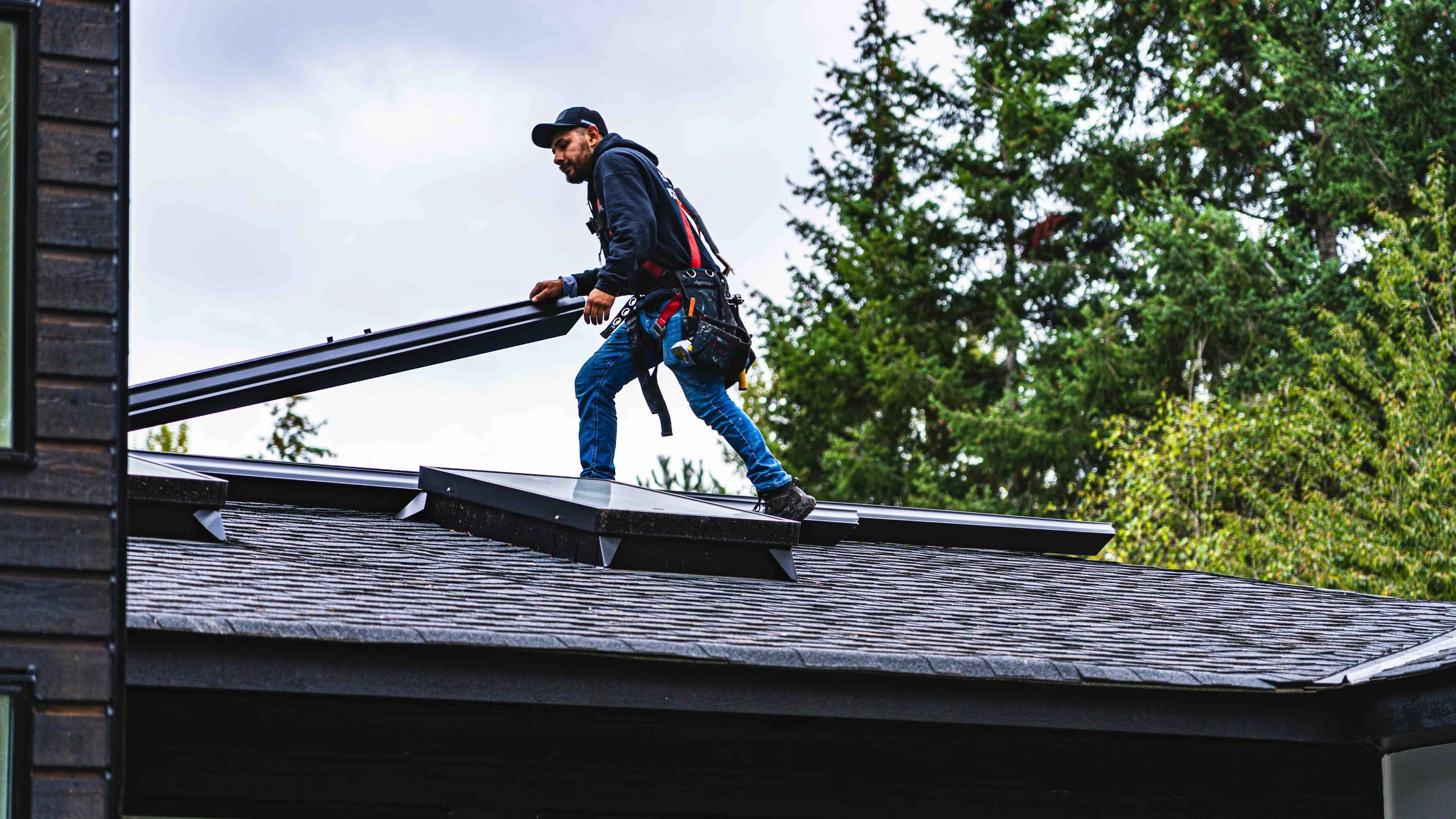 A man wearing a cap, hoodie, and work harness installing or repairing a metal roofing panel on a house.