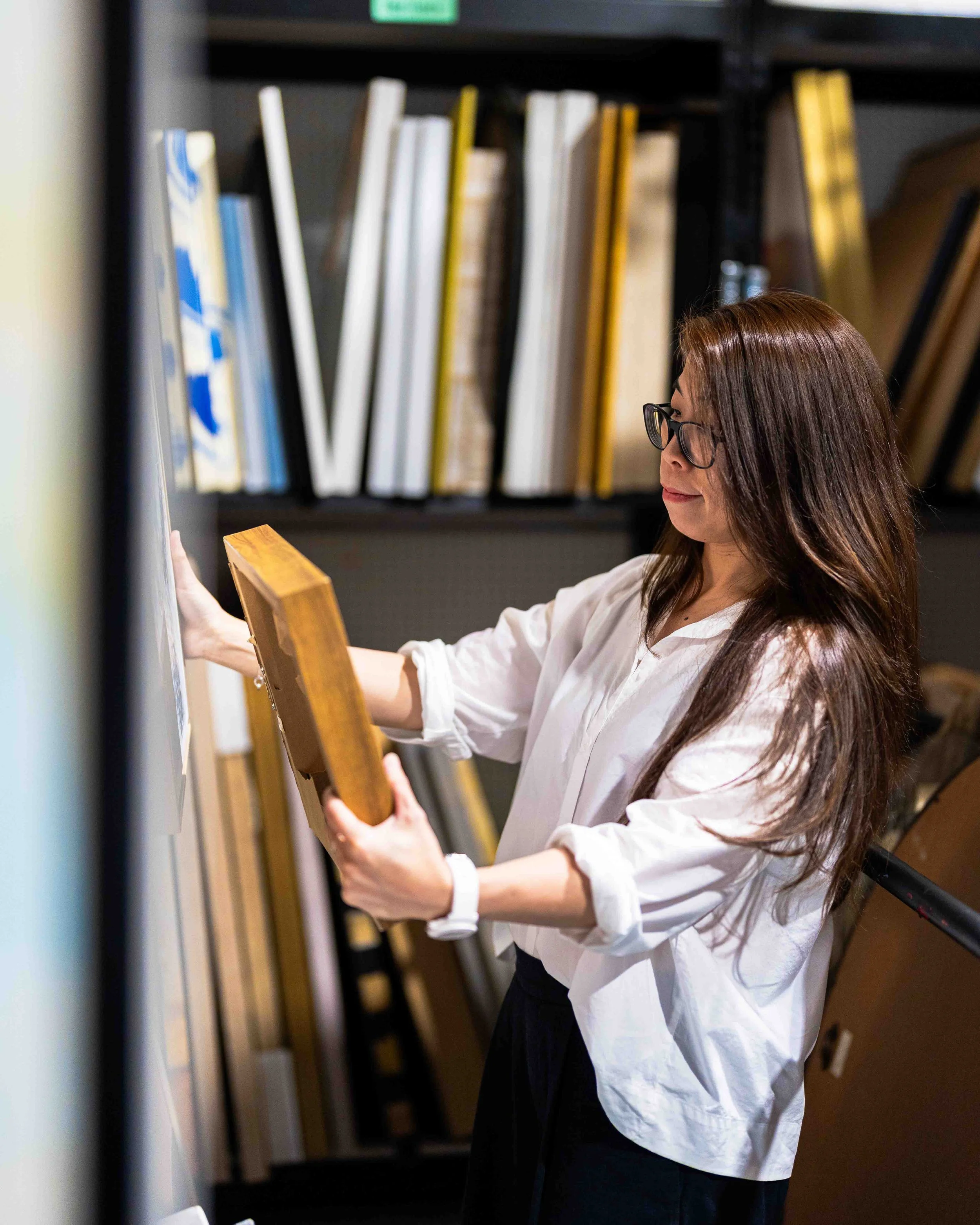 A woman with long brown hair and glasses in a white shirt is standing in a library or bookstore, looking at a book on a stand.
