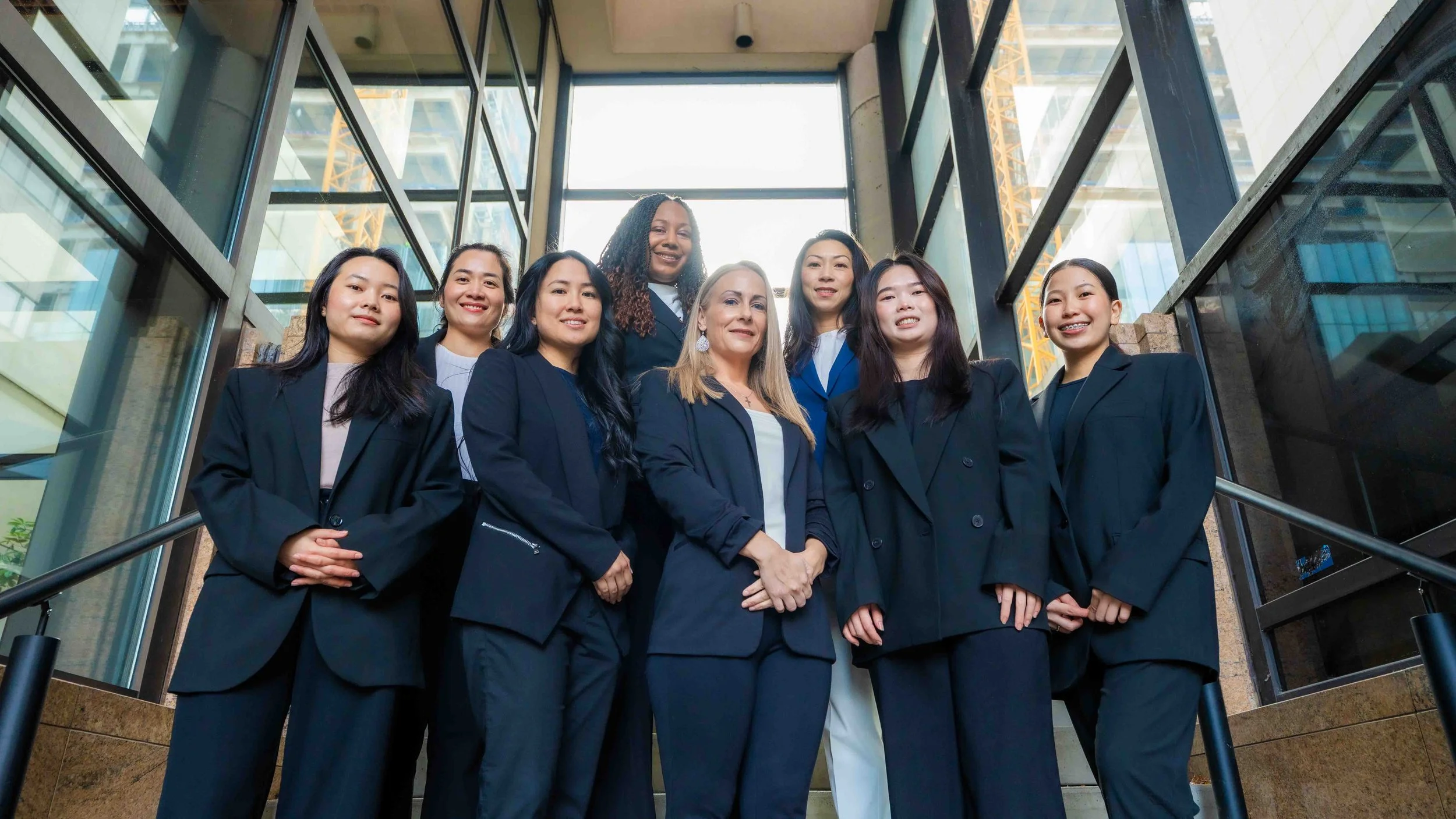 Group of diverse professional women standing together on a staircase inside a modern building with glass walls and a construction crane visible outside.