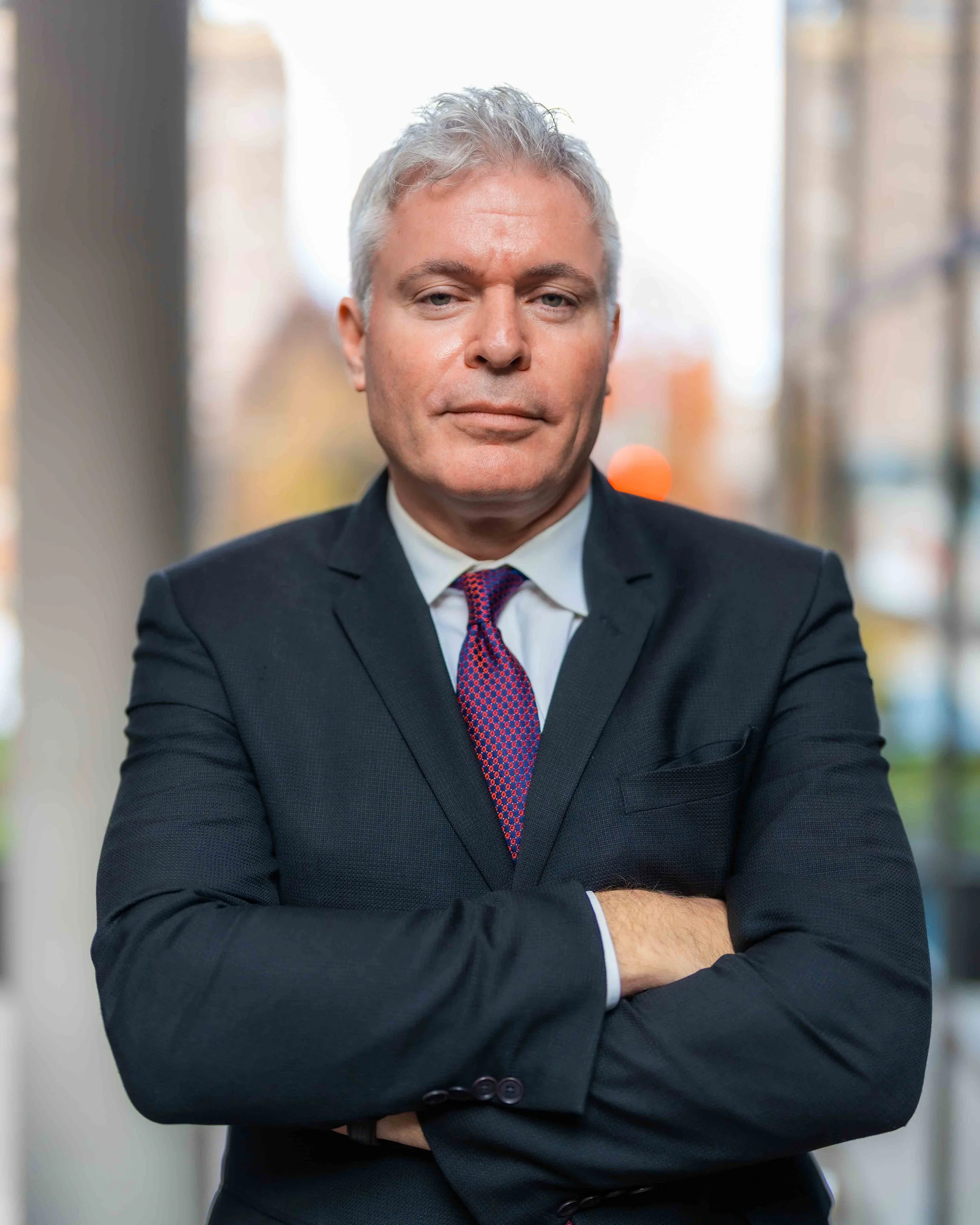 A middle-aged man with gray hair, wearing a dark suit, white shirt, and patterned red tie, standing outdoors with arms crossed, against a blurred background of buildings and trees.