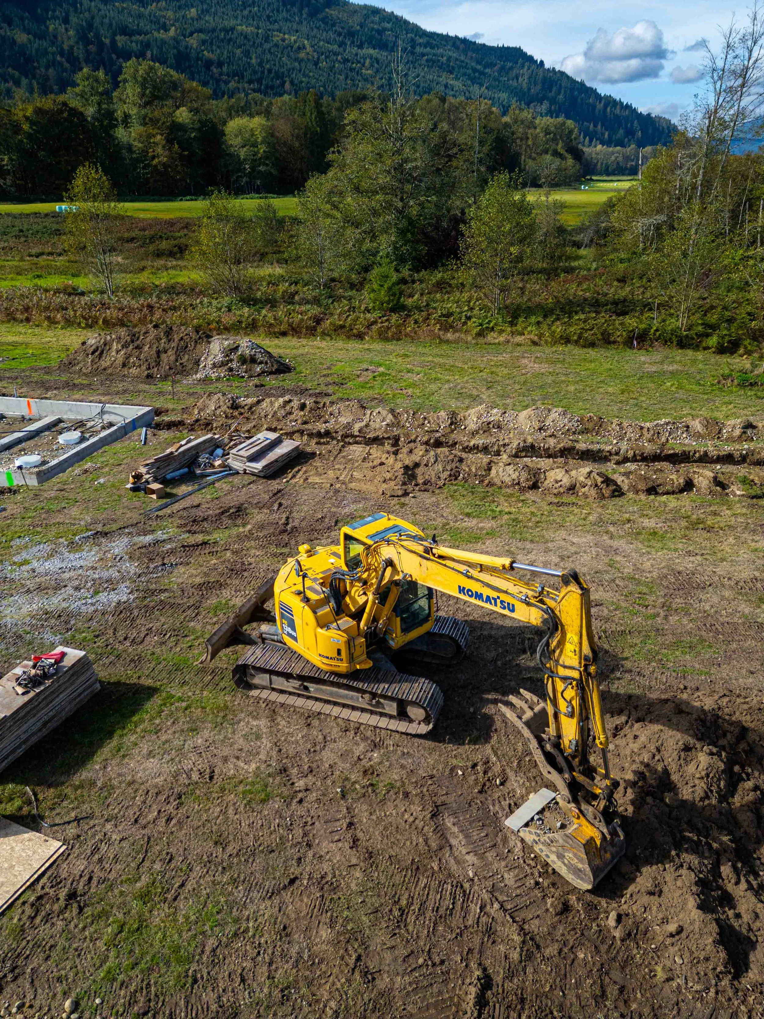 A yellow excavator working on a construction site with dirt and construction materials in the foreground, and trees, fields, and mountains in the background under a partly cloudy sky.
