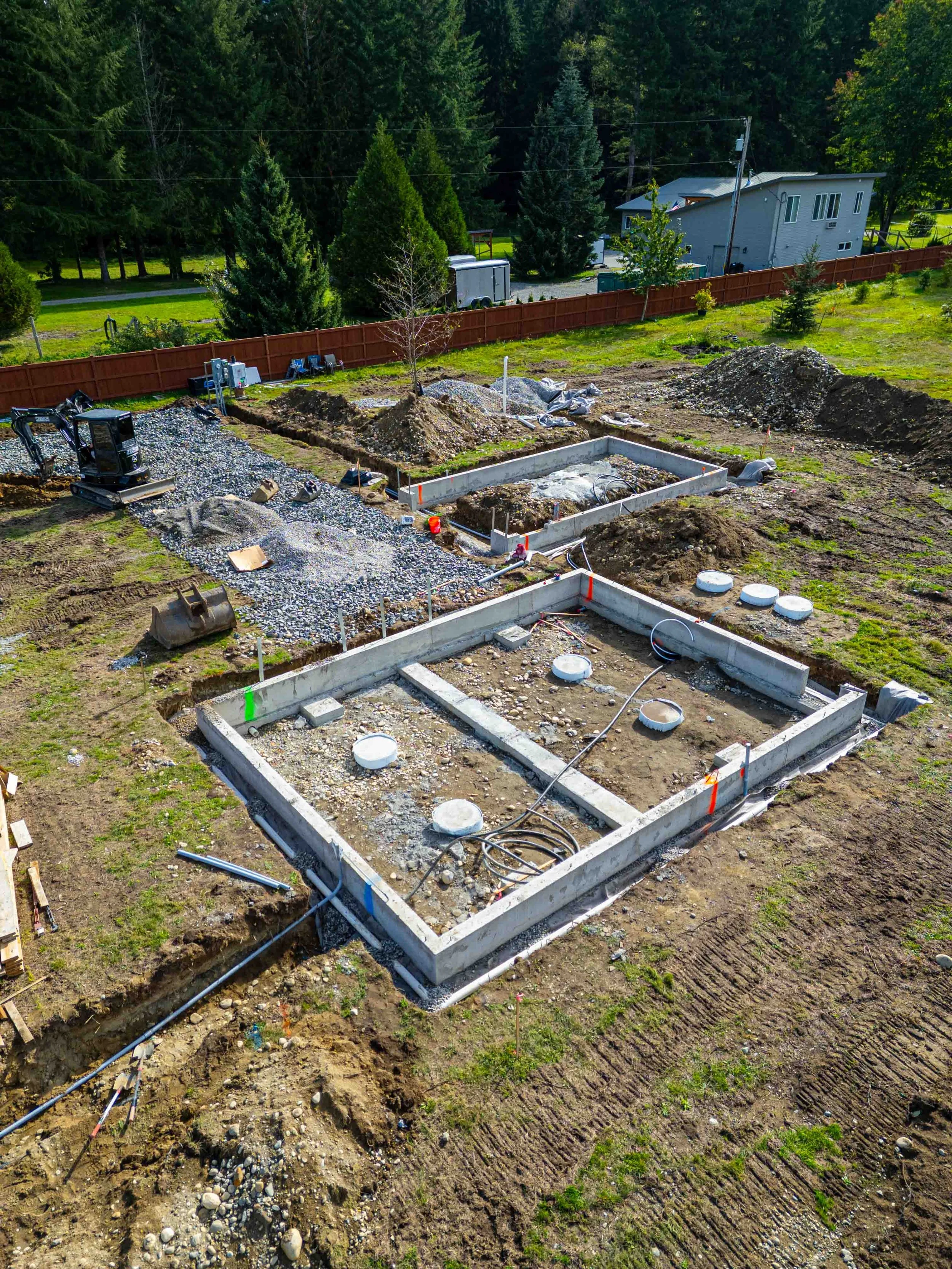 Construction site with foundation framework and gravel, surrounded by dirt and grass, with trees and a house in the background.
