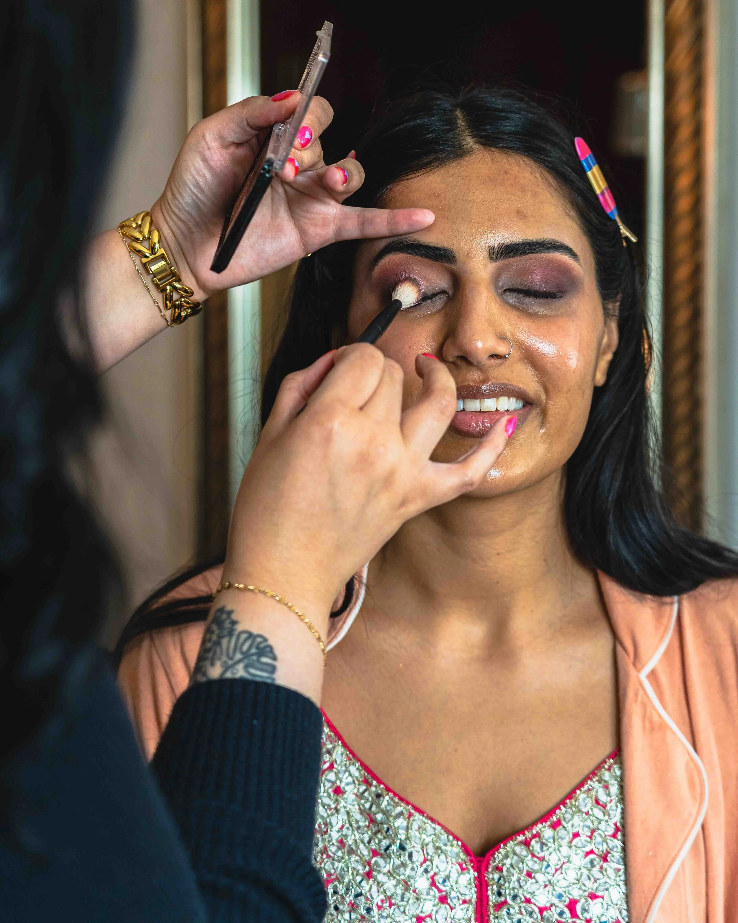 A woman with dark hair, wearing a coral-colored top with white embroidery and pink piping, is having her eyelid makeup done. A makeup artist with a gold bracelet and coral nail polish is applying eyeshadow with a brush. The woman is smiling with her 