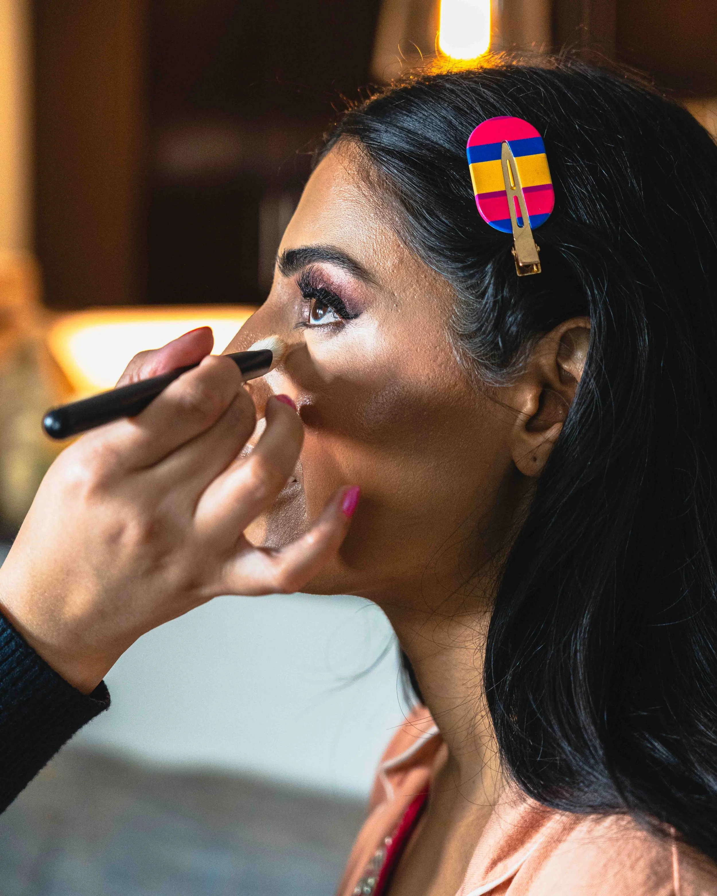 Close-up of a woman with dark hair wearing a colorful hair clip, applying makeup to her face with a brush, wearing a peach shirt with a gold chain necklace.