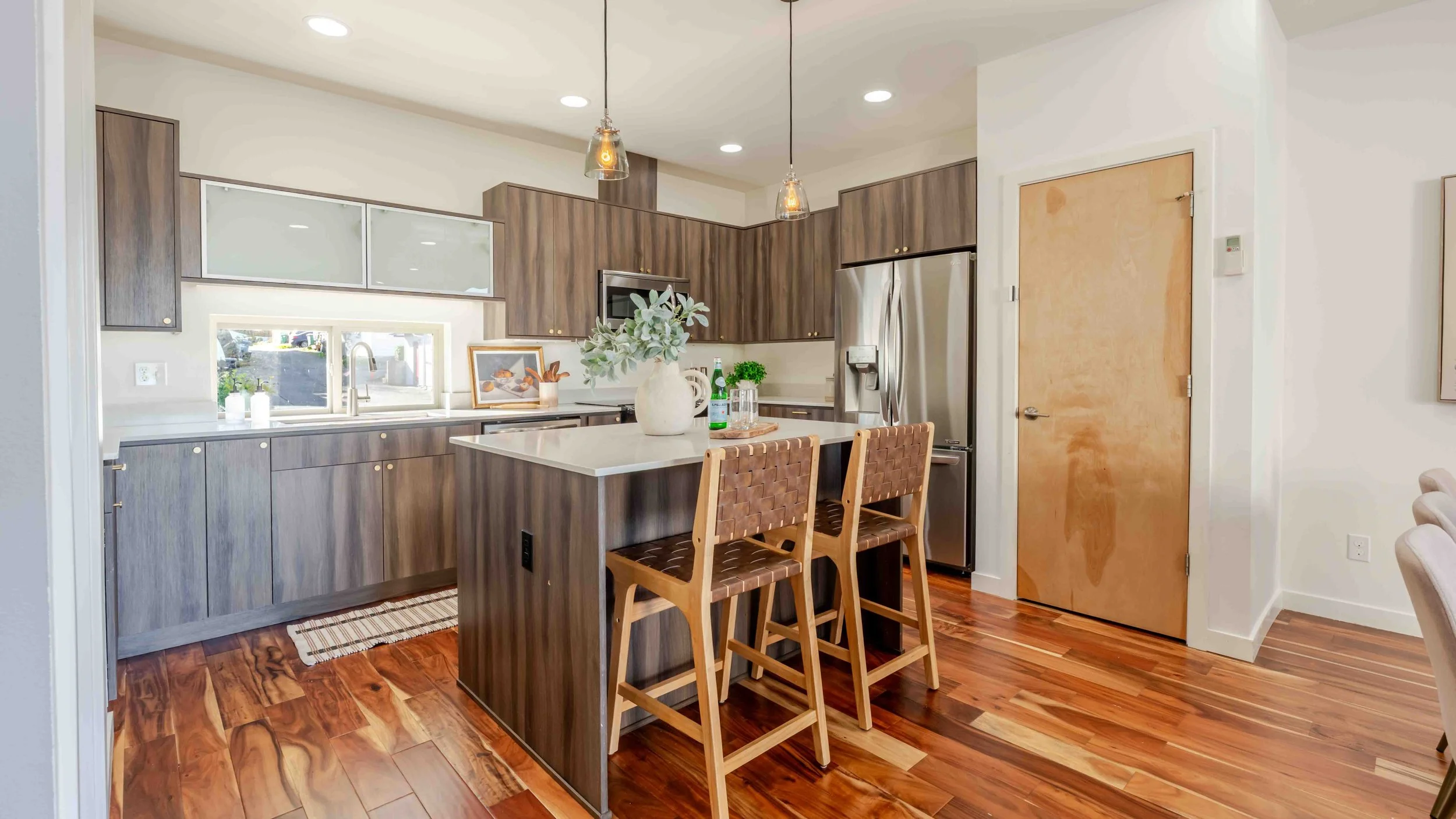 Modern kitchen with wooden cabinetry, a kitchen island with two woven leather chairs, stainless steel refrigerator, window above the sink, pendant lighting, and hardwood floors.