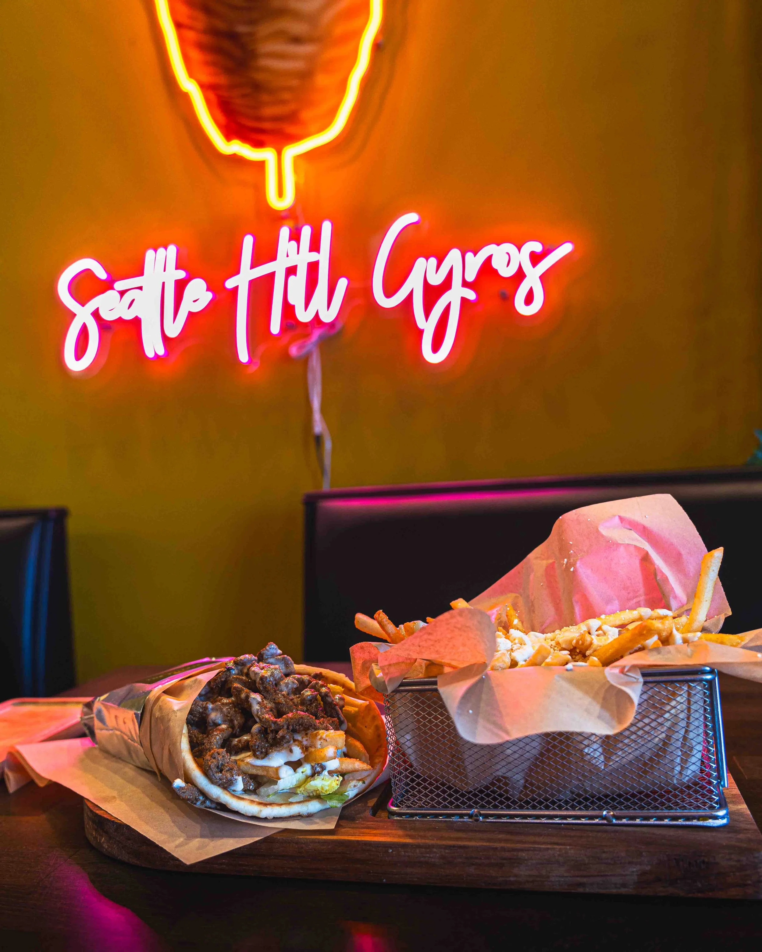 Neon sign reading 'Sit Still Gyros' in a dimly lit restaurant with two plates of gyros on the table, one with grilled meat and sauce, the other with fries and pita.
