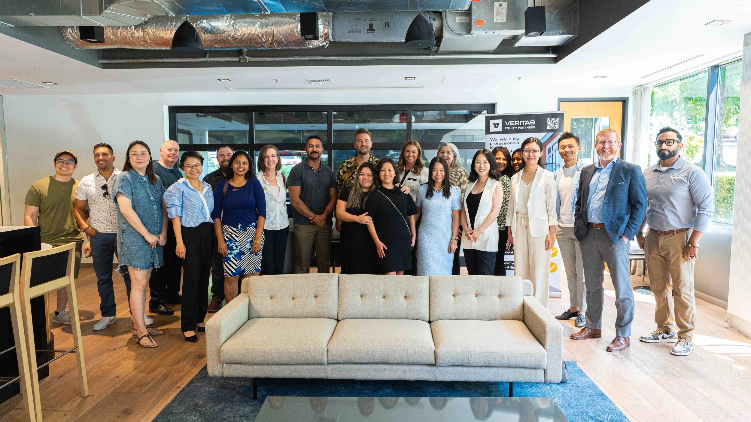 A large group of diverse people gathered indoors at a professional or social event, standing near a beige sofa, with a banner in the background indicating a corporate or organizational setting.