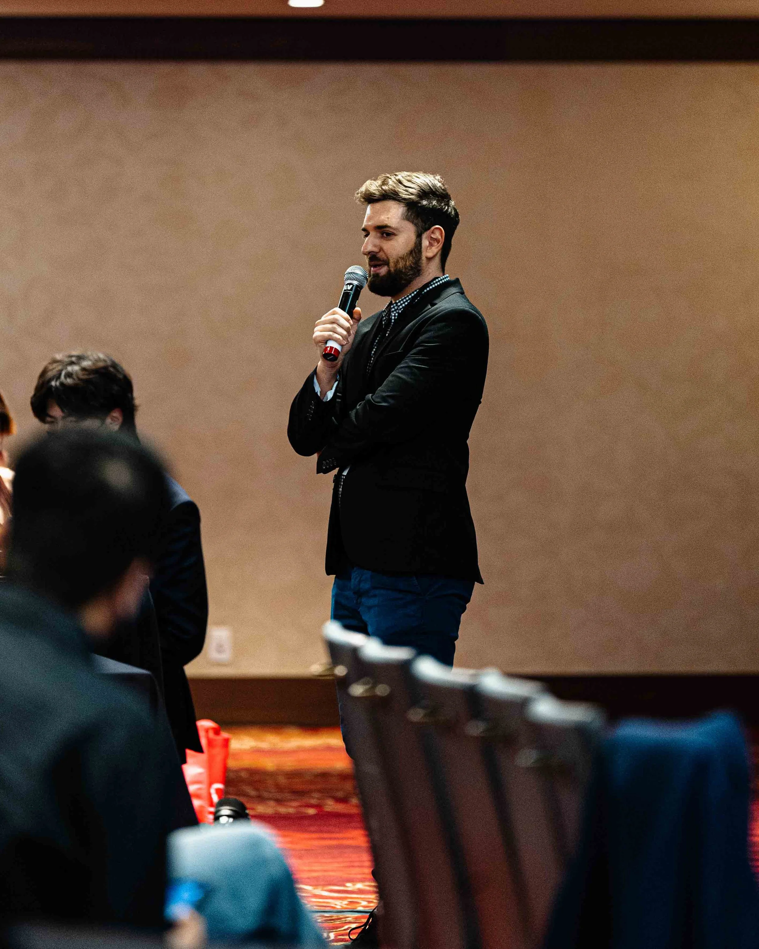 A man with dark hair and a beard is speaking into a microphone during an event or presentation, wearing a black blazer and a patterned shirt, standing in a room with a plain brown wall background.