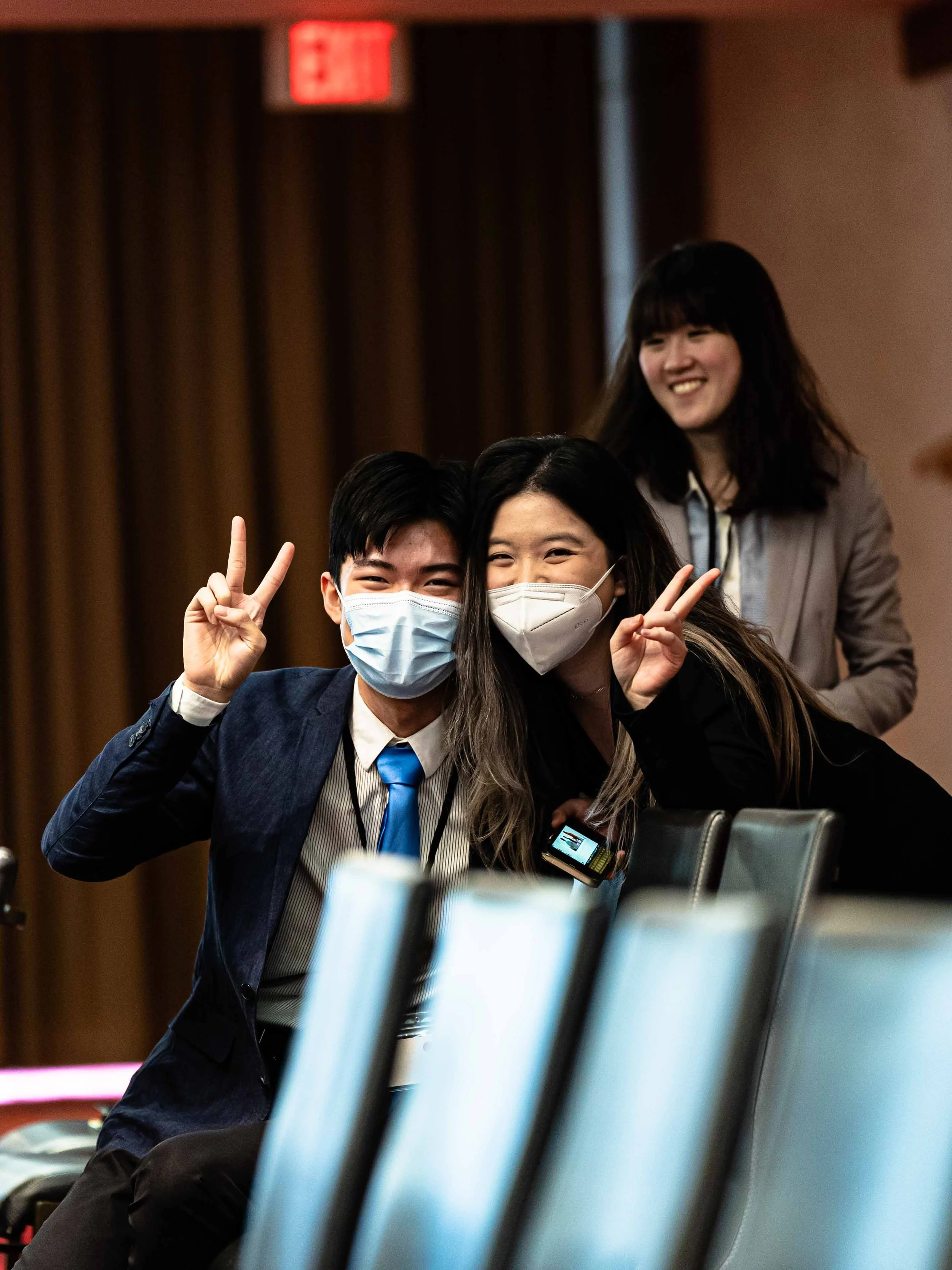 Two people wearing face masks posing for a photo, making peace signs with their fingers. They are seated in a room with a woman standing behind them, smiling. The background has a red exit sign and brown curtains.