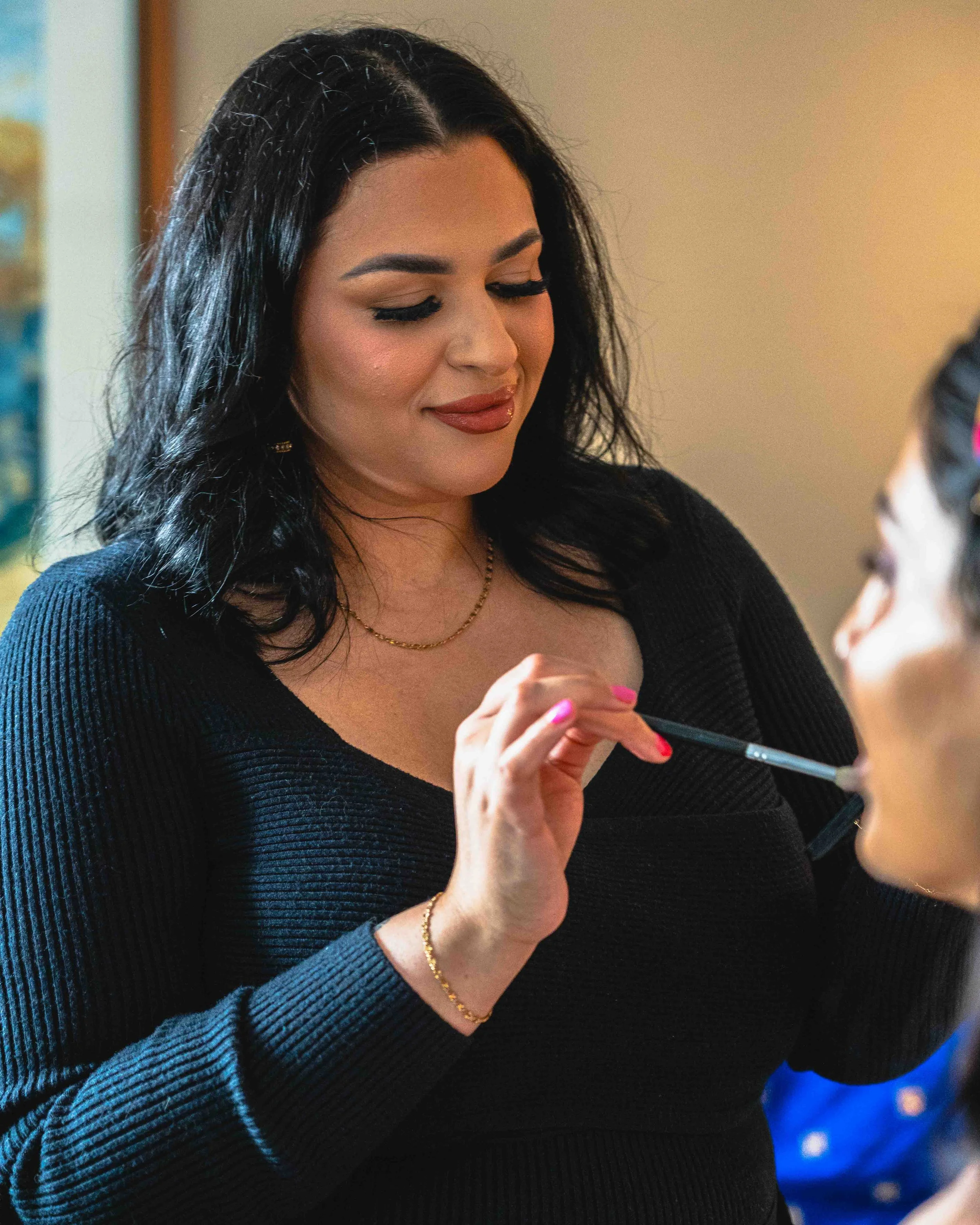 A woman with dark, curly hair, wearing a black ribbed sweater, gold necklace, and gold bracelet, is smiling and holding a makeup brush. She appears to be looking at her reflection in a mirror.