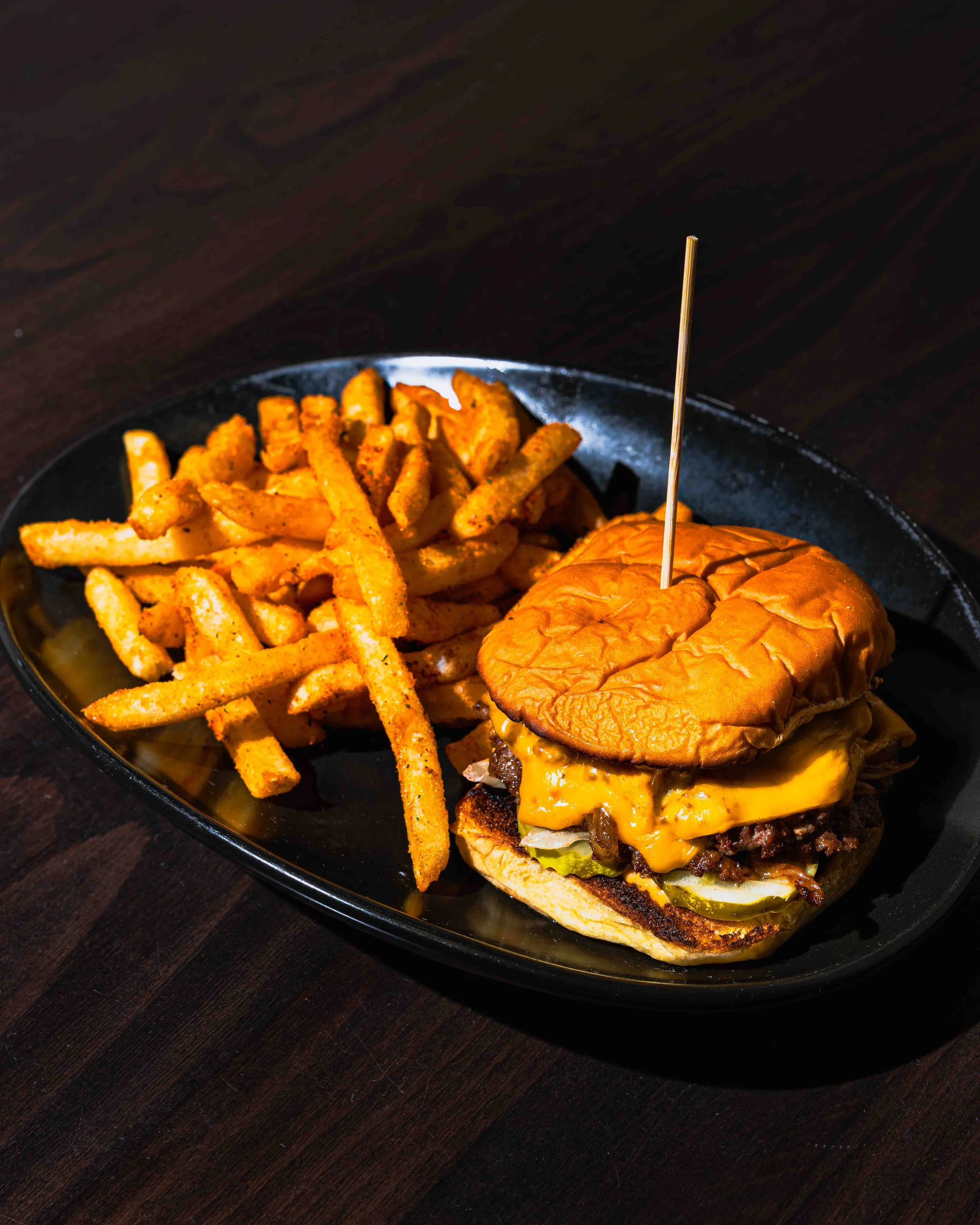 Close-up of a cheeseburger with lettuce, tomato, melted cheese, and a bun on a black plate, accompanied by seasoned French fries, on a dark wooden surface.