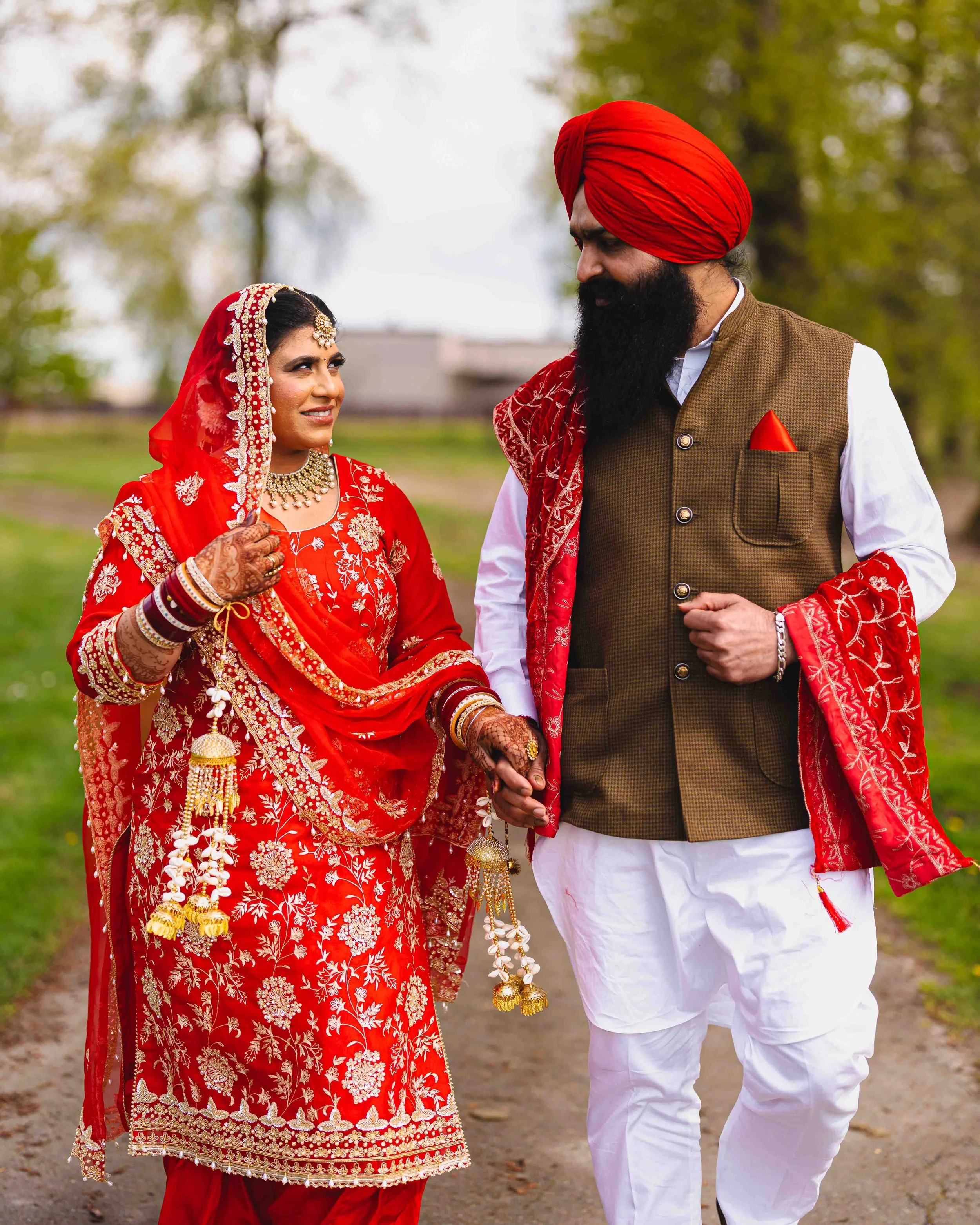 A couple in traditional Indian wedding attire holding hands outdoors, with the woman in a red and gold embroidered saree and the man in a brown vest, white kurta, and red turban, walking on a path with trees in the background.