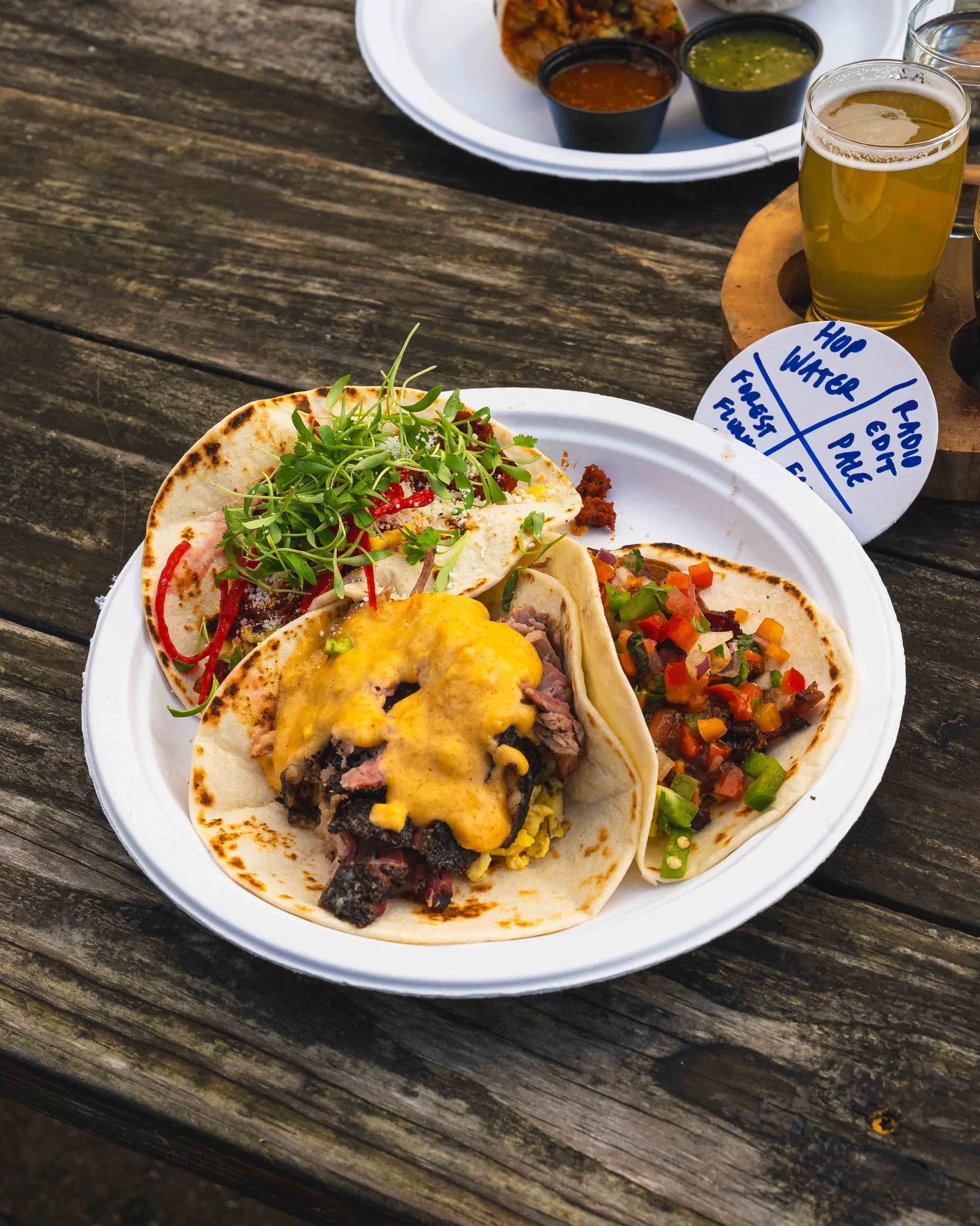 Close-up image of a plate with four tacos filled with various ingredients, topped with sunflower microgreens, on a weathered wooden table. In the background, there is a white plate with chips and three small containers of dipping sauces, along with t