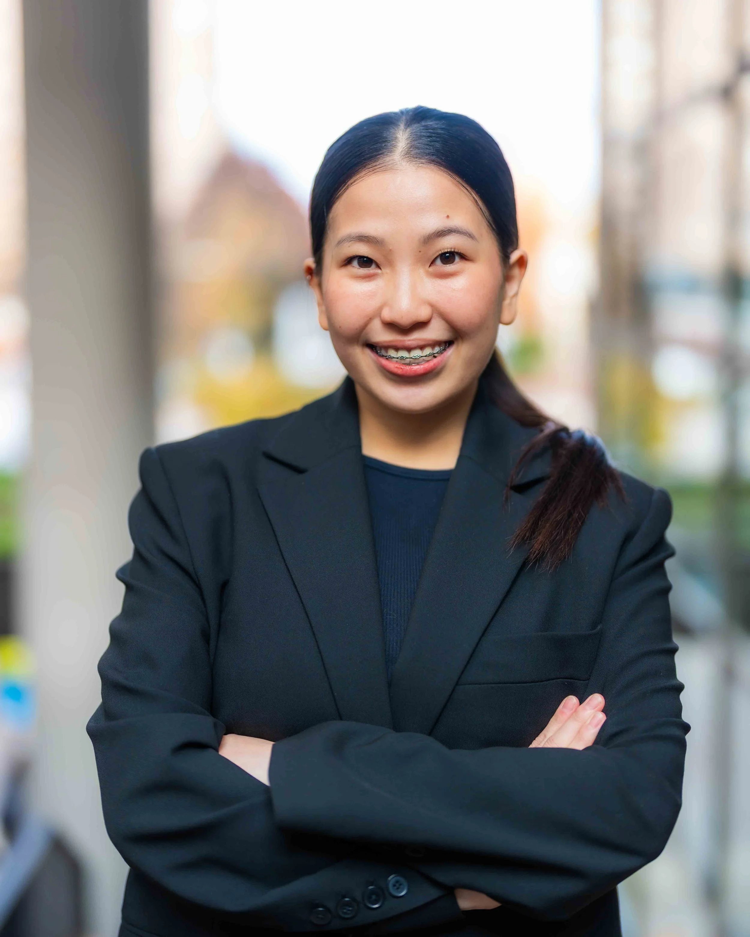 A woman in a black blazer smiling with braces, arms crossed, standing outdoors in a blurred urban background.
