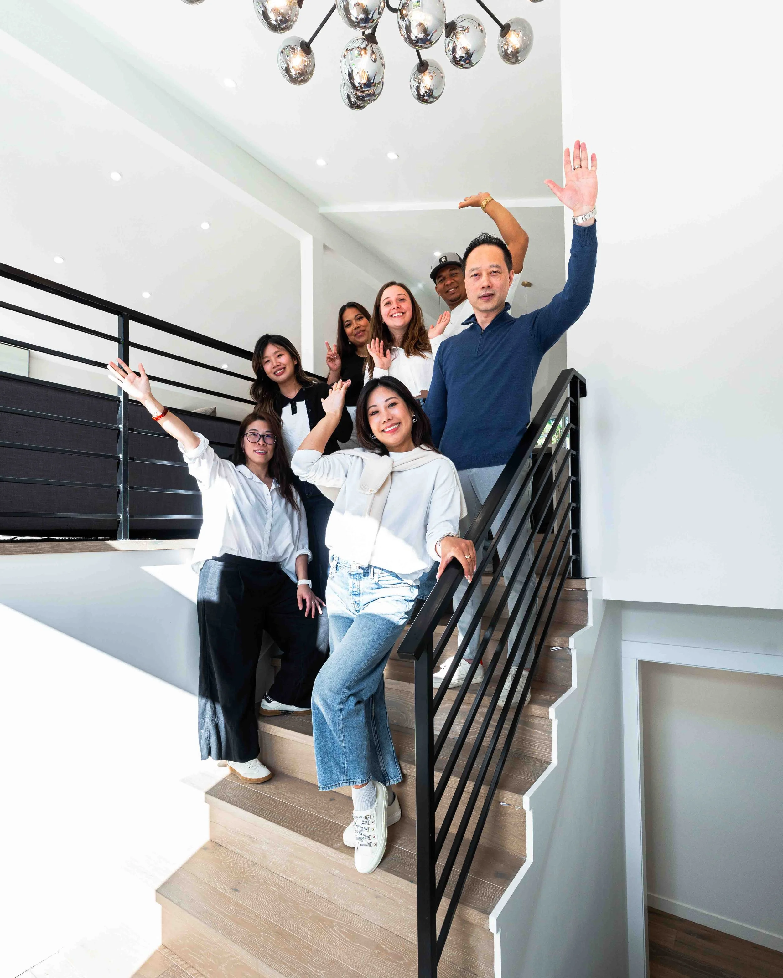 A diverse group of seven people standing on a staircase, waving and smiling at the camera in a modern, bright interior with white walls and a metallic chandelier hanging from the ceiling.