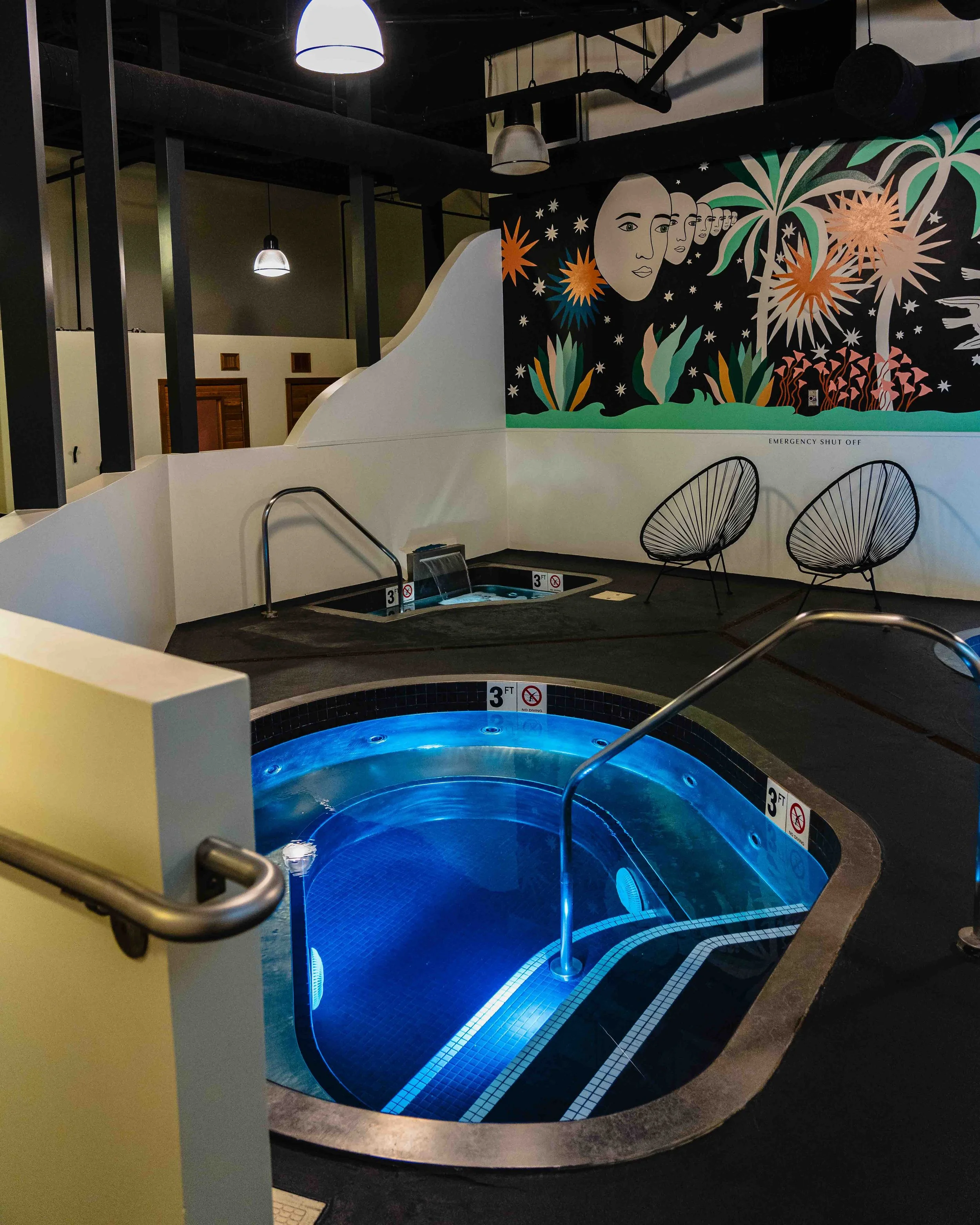 Indoor hot tub with blue glowing water, metal handrails, and an adjacent small pool with similar lighting. The background features a black wall with a colorful mural depicting a woman's face, stars, moons, and tropical plants, in a modern, artistic s