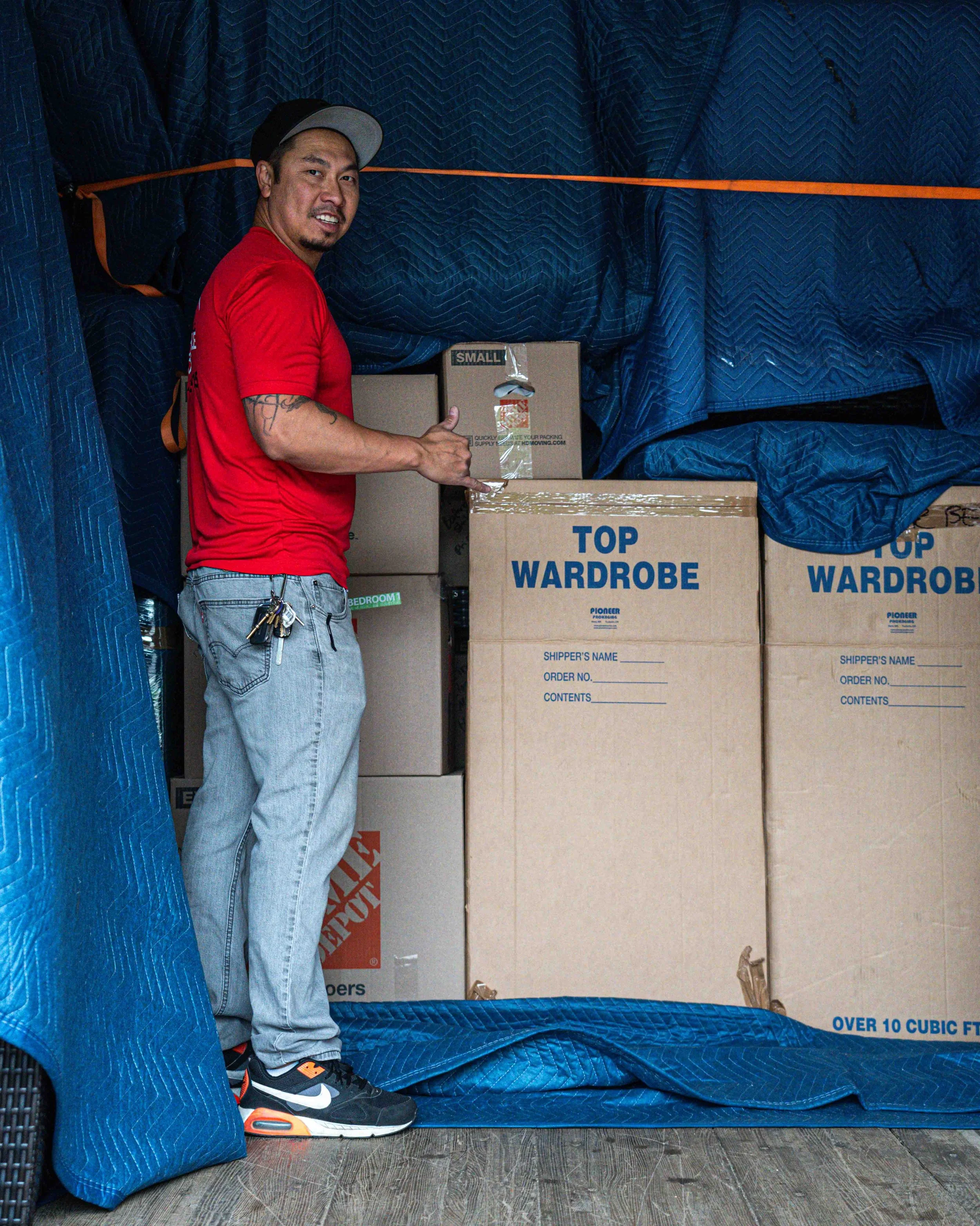 Man in red shirt and gray jeans standing inside a storage area surrounded by moving boxes wrapped in blue moving blankets.