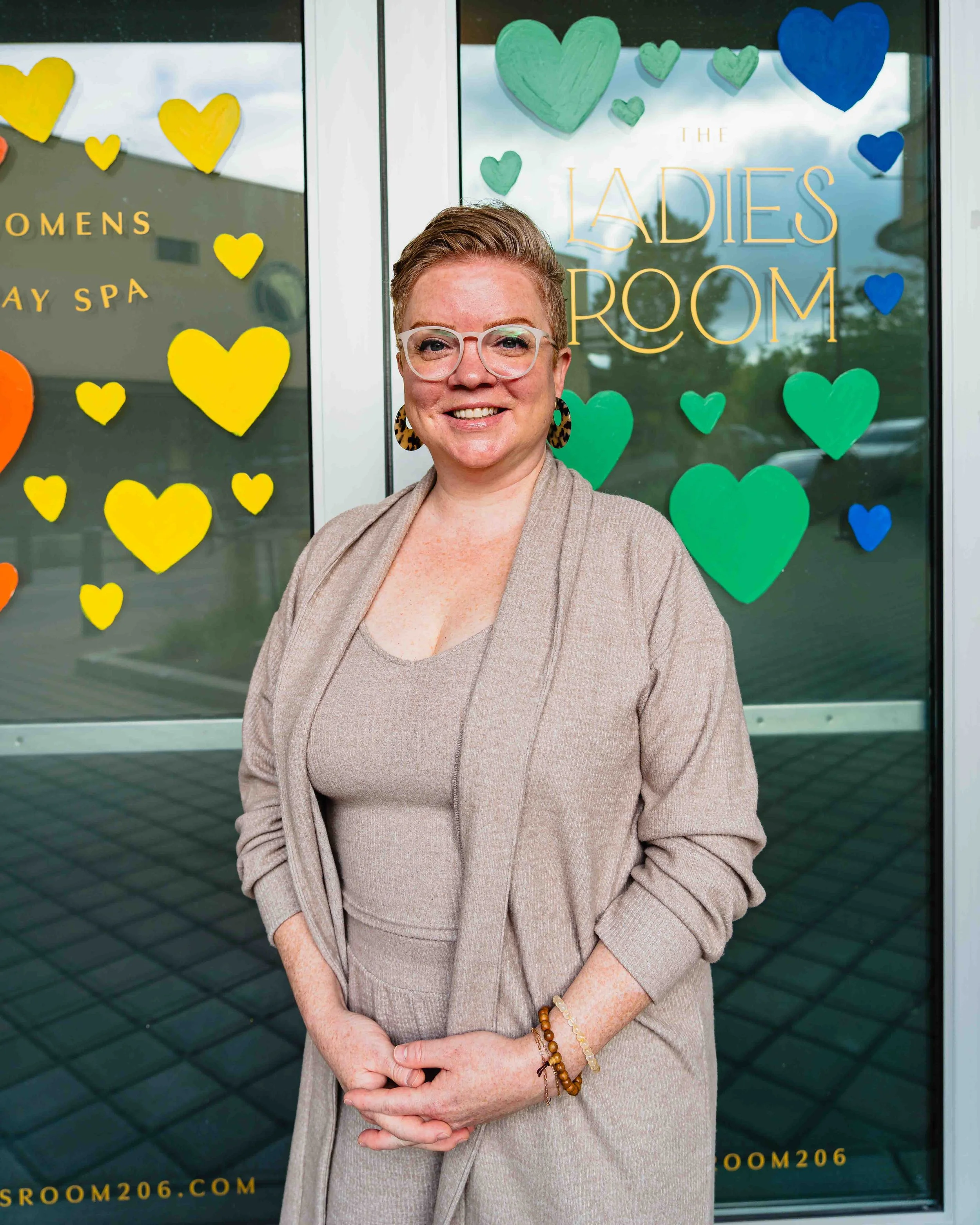A smiling woman with short red hair, glasses, and leopard print earrings, standing outside near a window with colorful paper hearts and signs reading "The Ladies Room" and "Women".