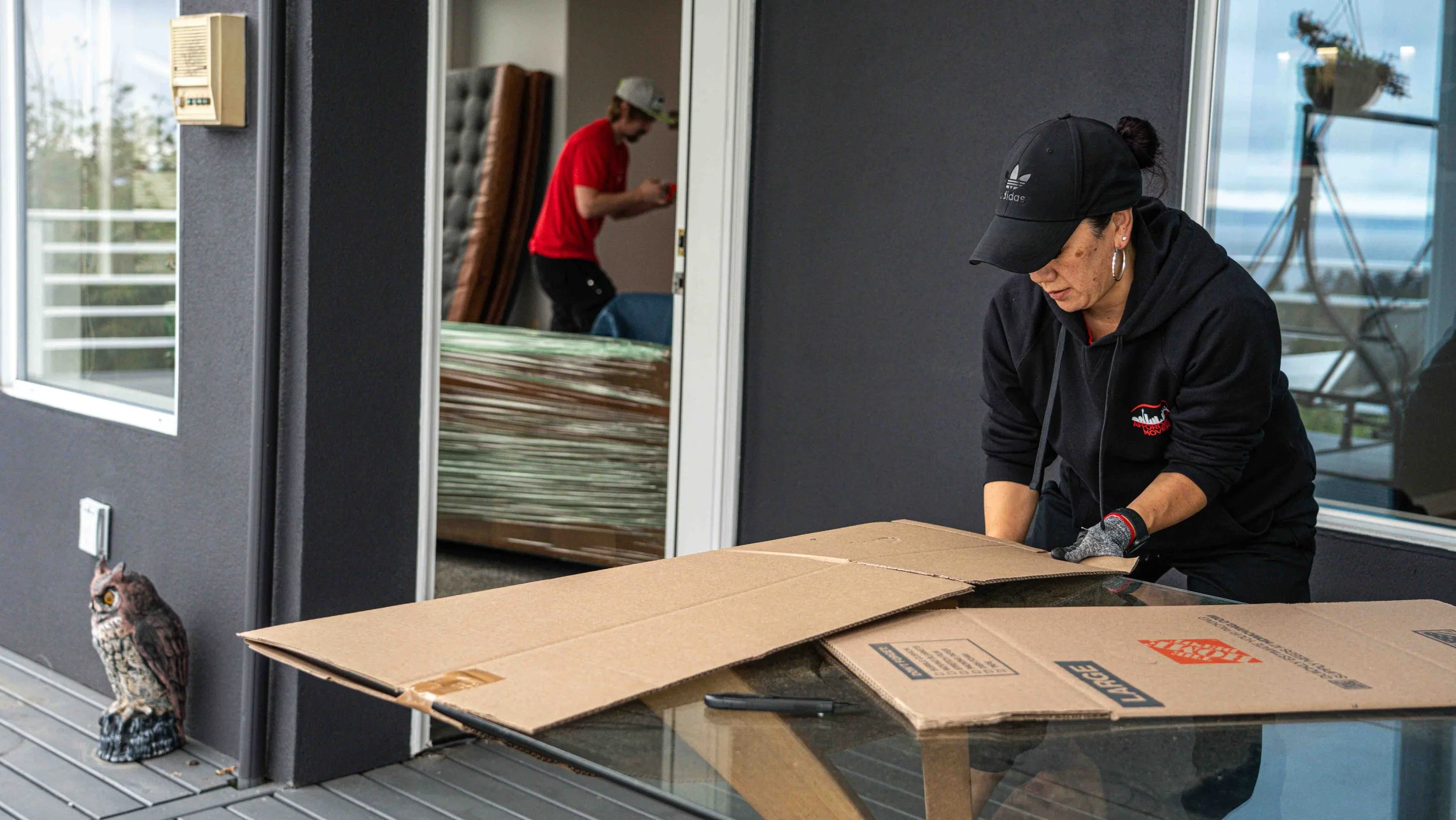 A woman with a black cap and hoodie is bending over a table, taping a cardboard box. A gray owl figurine is on the porch, and in the background, a man in a red shirt and gray cap is handling moving boxes near the doorway.