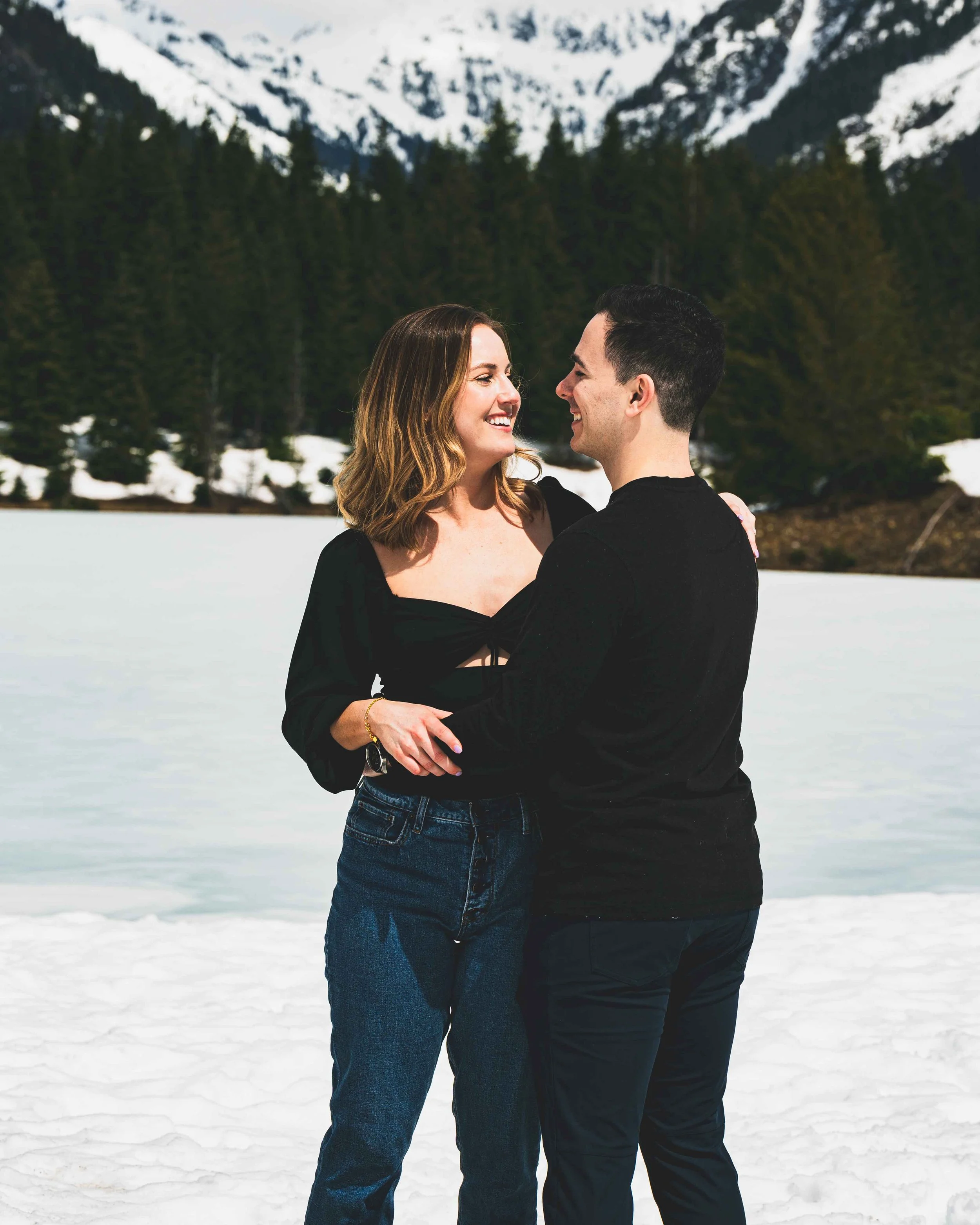 A smiling couple facing each other and holding each other in a snowy outdoor setting with mountains, evergreen trees, and snow-covered terrain in the background.