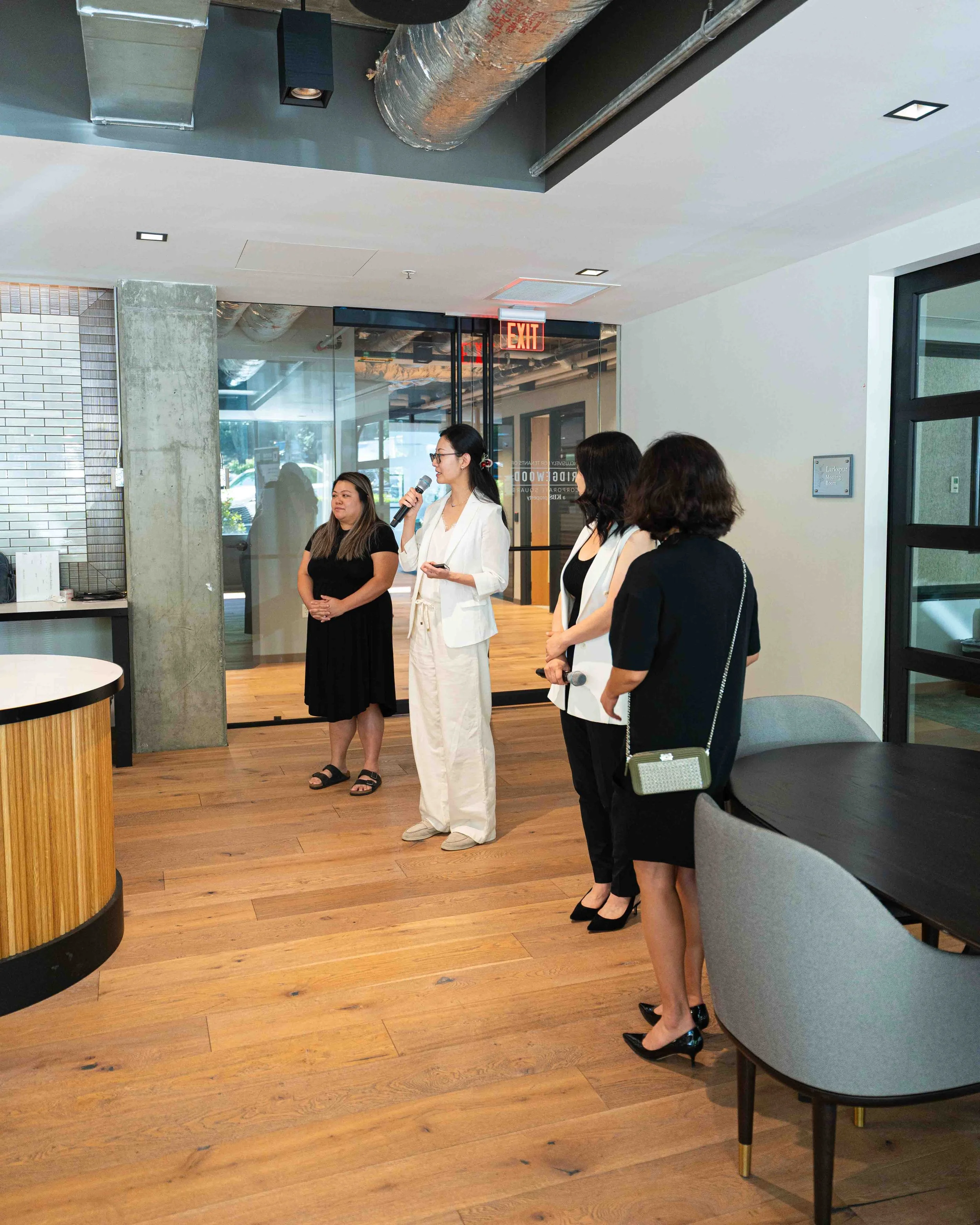 Group of women standing in a modern office space, one woman in a white suit speaking into a microphone, others listening attentively, with a mix of casual and professional attire, in a room with wooden flooring, contemporary chairs, and industrial ce