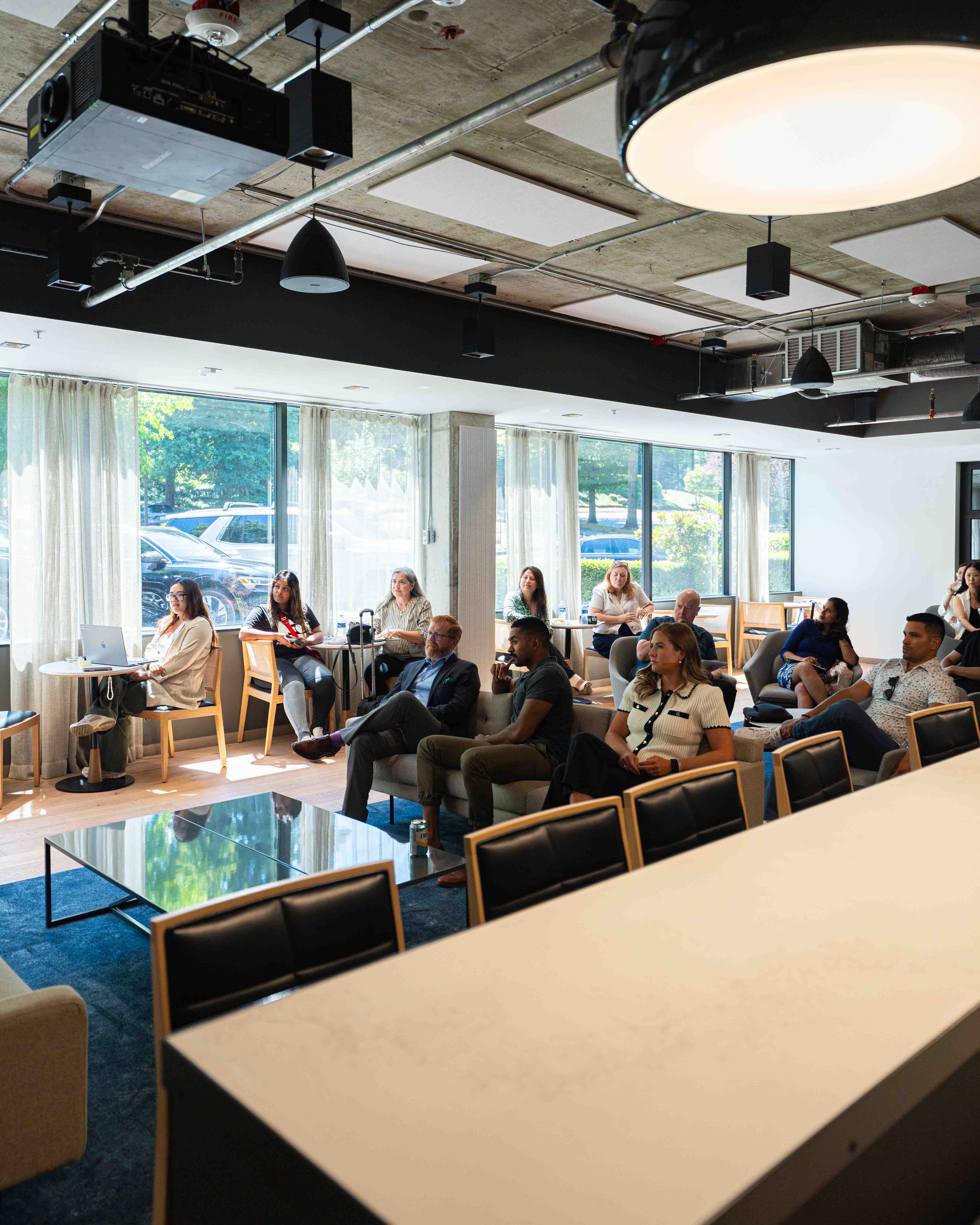 A group of people sitting in a modern, well-lit room with large windows. Some are using laptops, and others are listening attentively, suggesting a meeting or workshop setting. The room features a concrete ceiling with exposed pipes and ceiling-mount