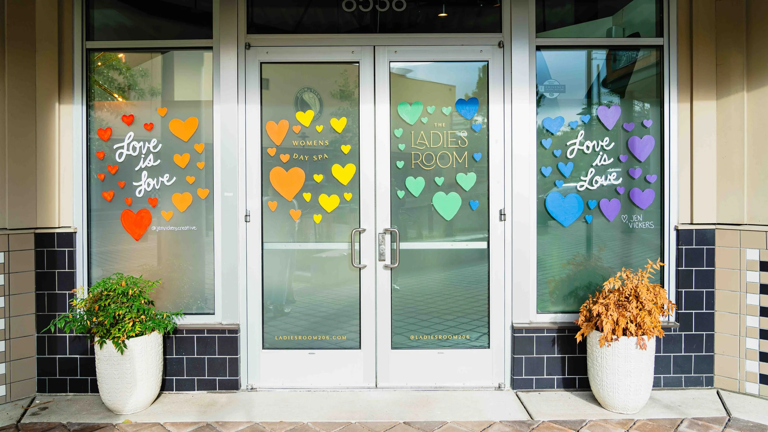 Storefront with glass doors decorated with colorful paper hearts and signs, including 'Love is Love,' 'Womens Day Spa,' and 'The Ladies Room.' Potted plants with autumn leaves flanking the entrance.