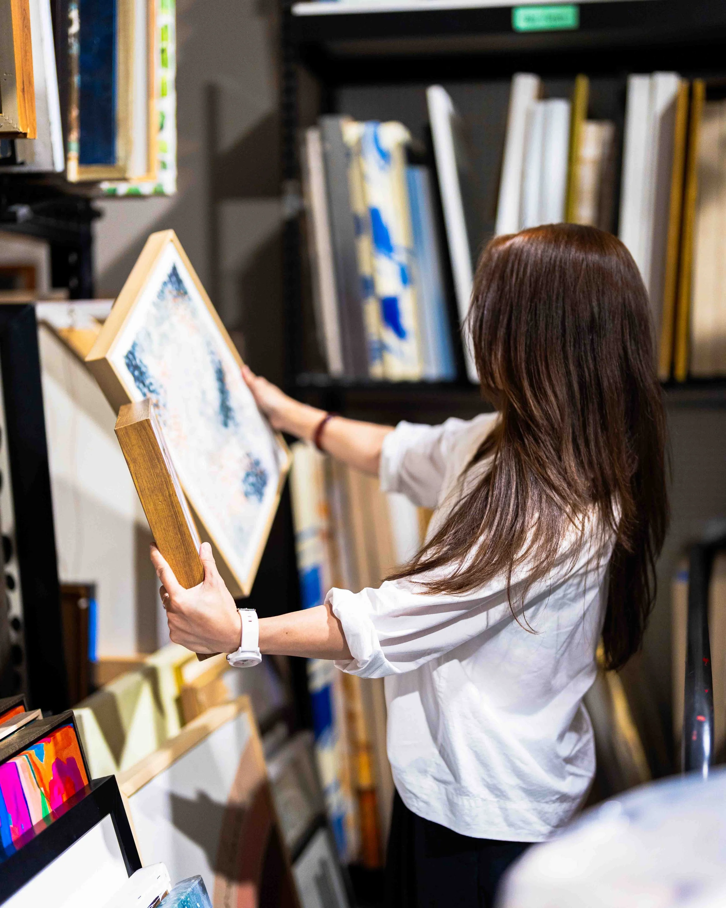 A person with long brown hair in a white shirt is looking at artwork in a gallery or bookstore, holding a framed piece of art. The background shows shelves filled with books and artwork.