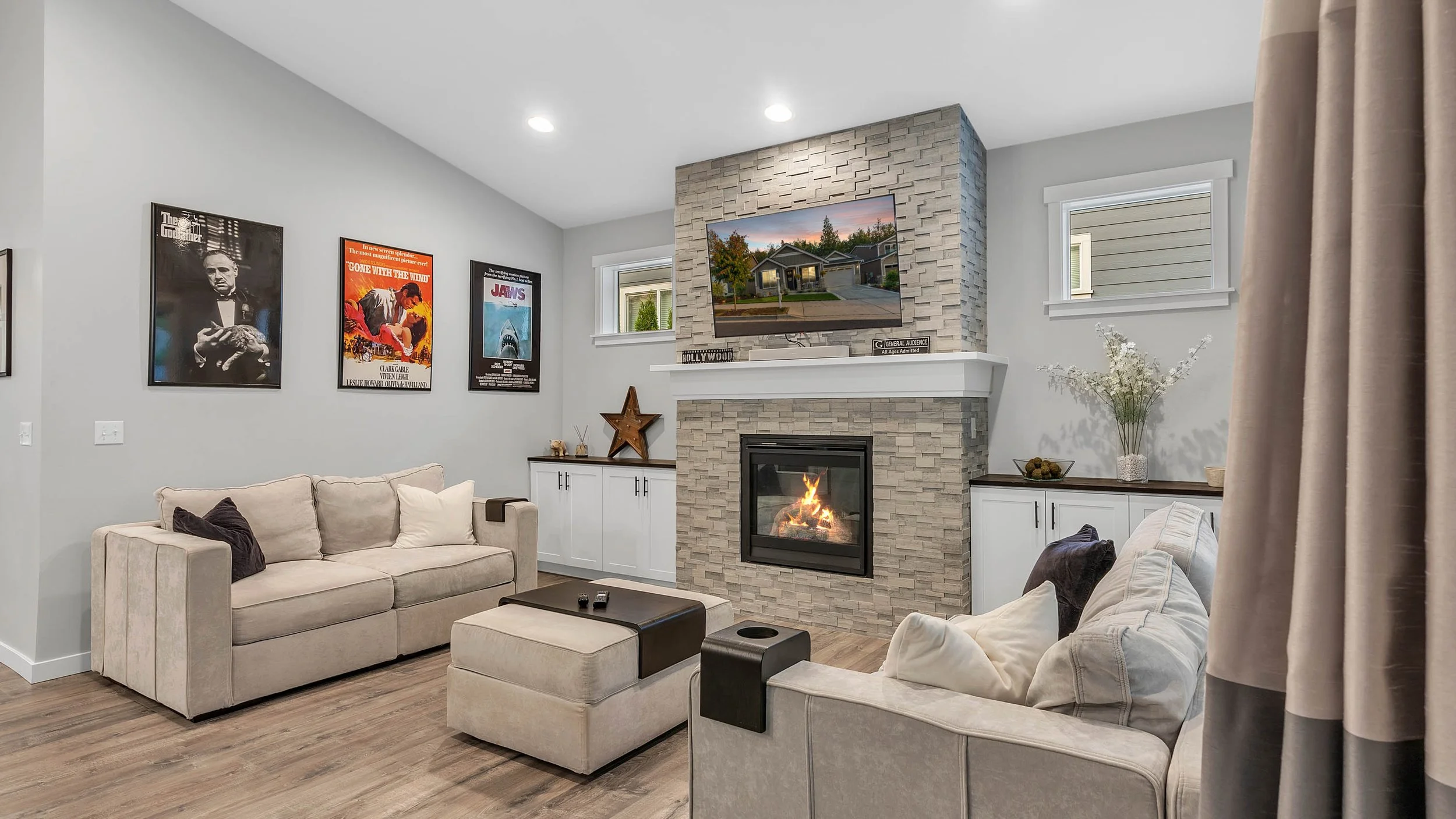 A modern living room with a beige sofa, white ottoman, and a fireplace with a stone surround. Wall art includes framed posters, and the décor features a star ornament, vases, and flowers. A flat-screen TV is mounted above the fireplace, showing a hou