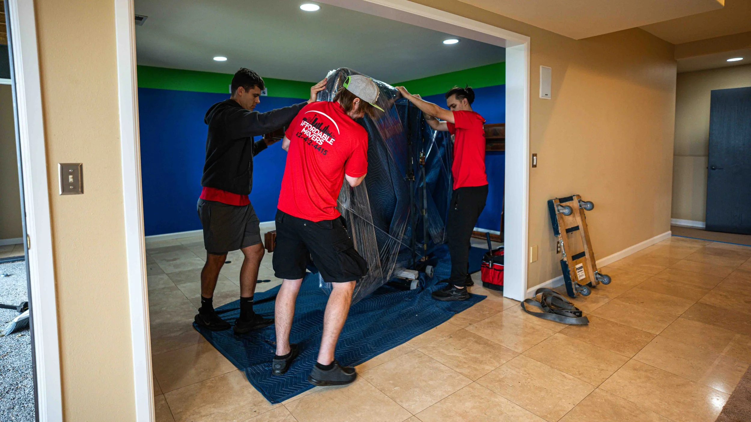 Four men are moving a large, wrapped piece of furniture into a house. They are working together, some holding the furniture and others guiding it.