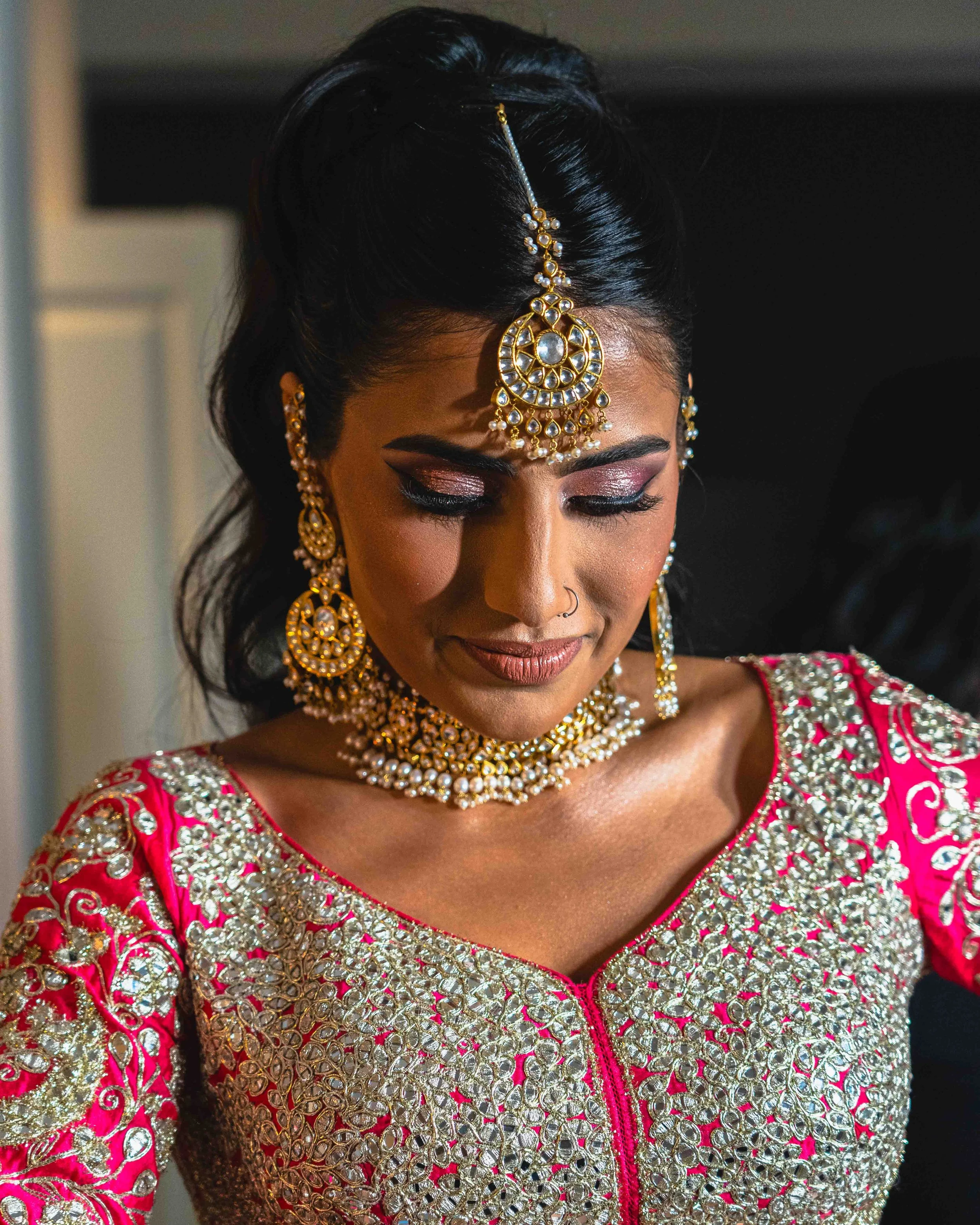 Close-up of a woman dressed in traditional Indian attire with elaborate gold and white jewelry, including a maang tikka on her forehead, earrings, and layered necklaces, wearing a bright pink and silver embroidered outfit.