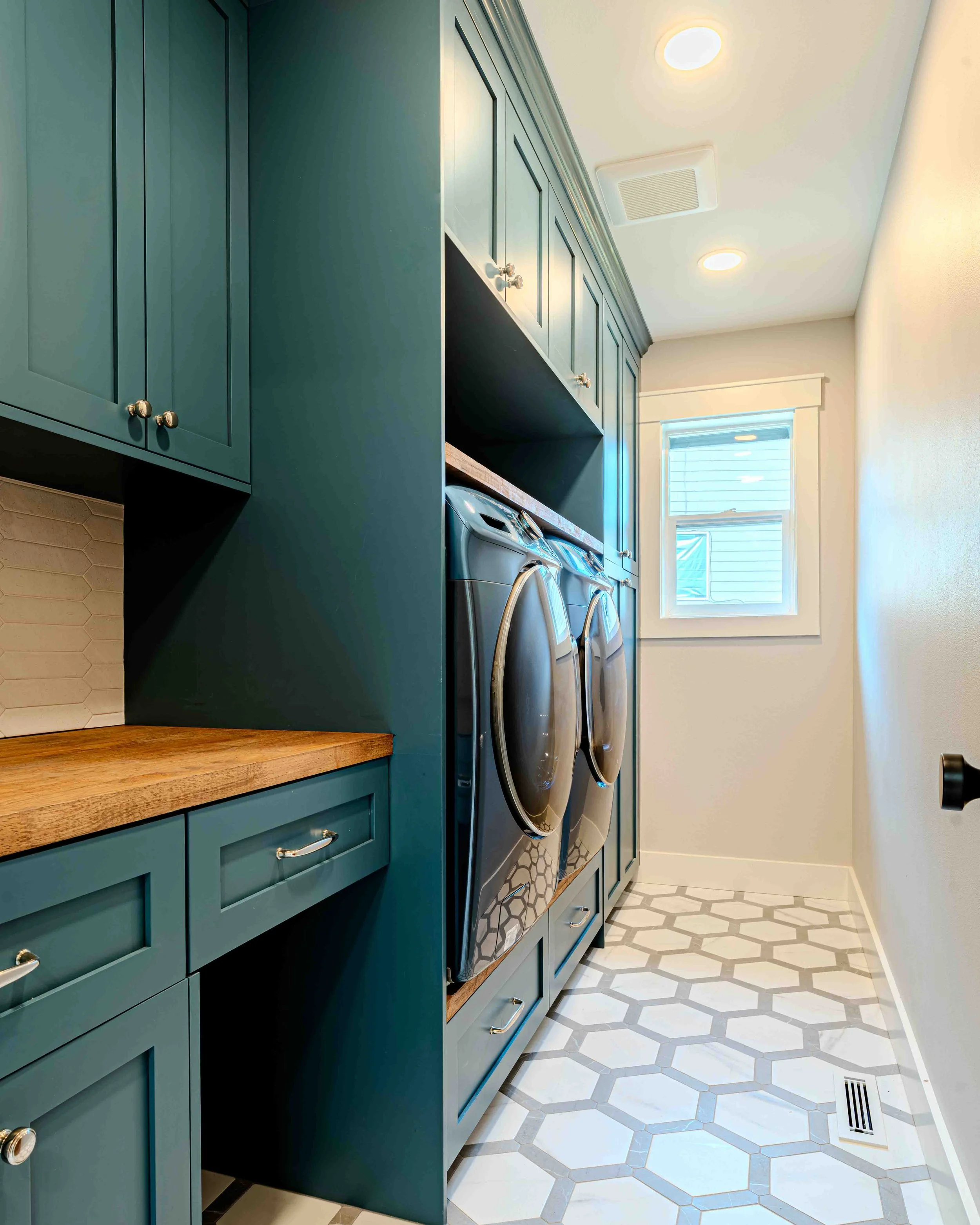 A laundry room with teal cabinets, wooden countertop, front-loading washing machines, and hexagonal tiling on the floor. White window with trim, ceiling lights, and an air vent are also visible.