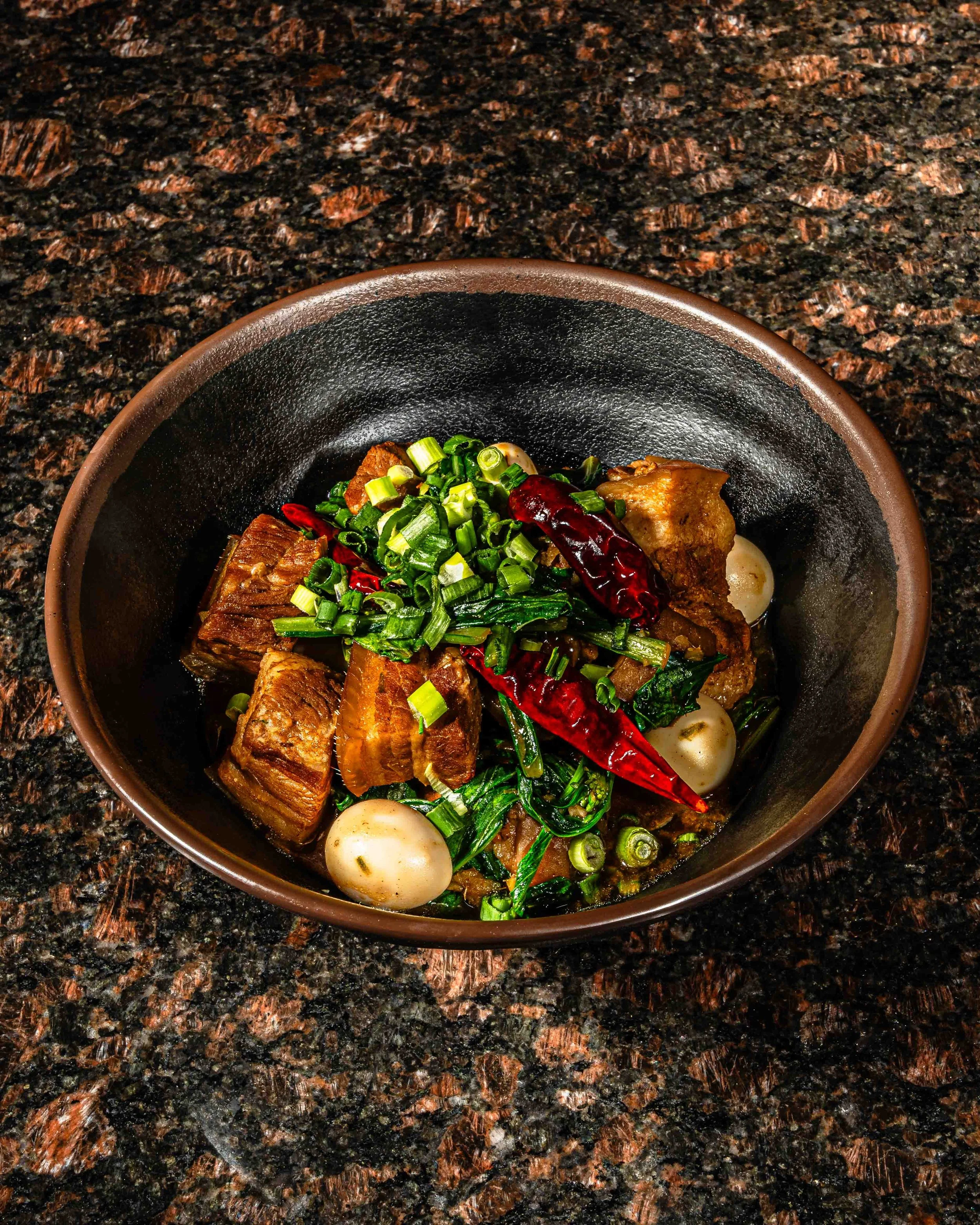 A bowl of Asian-style braised pork belly with boiled eggs, green onions, and dried red chili peppers on a dark granite countertop.