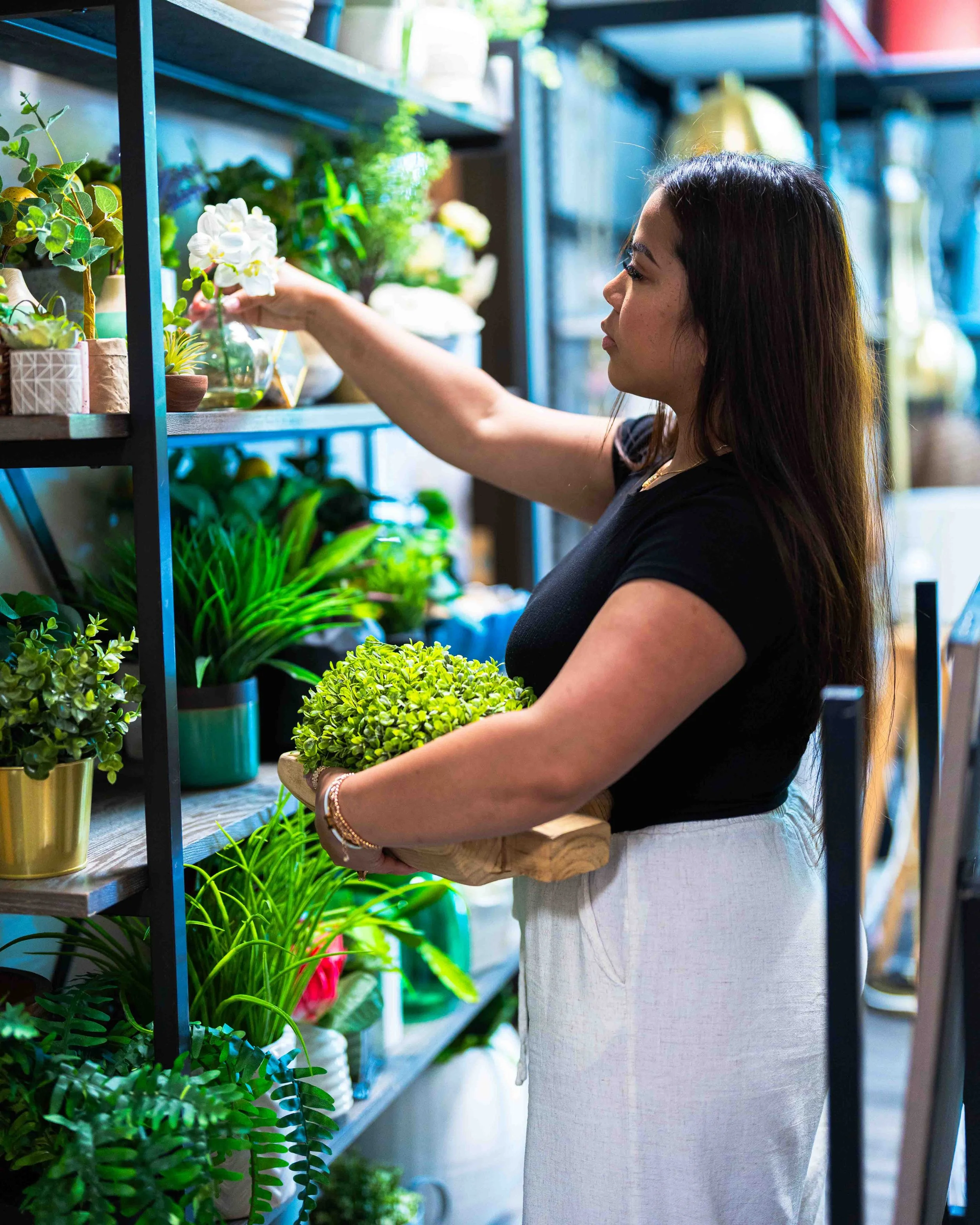 A woman in a black shirt and white linen pants is shopping for plants and flowers in a store. She is reaching for a white flower with her right hand and holding a small pot with green plants in her left hand. The store has metal shelves filled with p