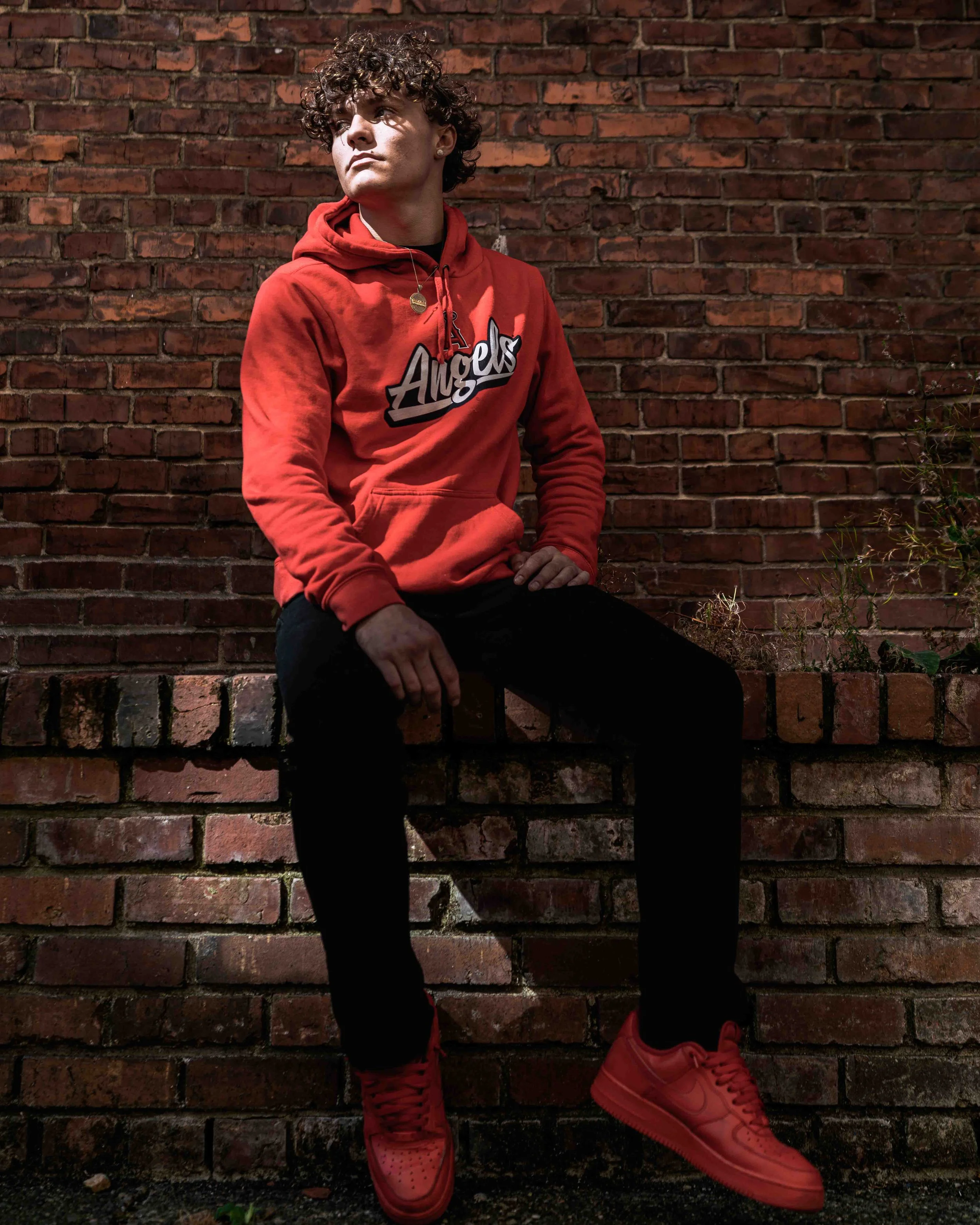 A young person with curly hair wearing a red hoodie with the word 'Angels' on it, sitting against a brick wall, with one foot resting on a bench or ledge and the other extended outward, wearing red sneakers.