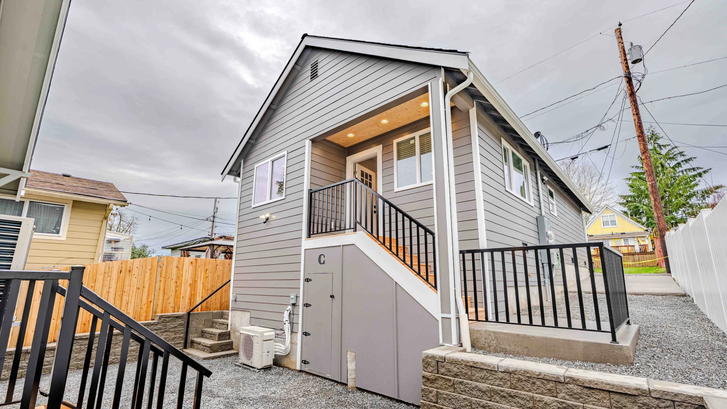 A modern gray house with a front porch and stairs, surrounded by a gravel driveway, a wooden fence, and neighboring houses under a cloudy sky.