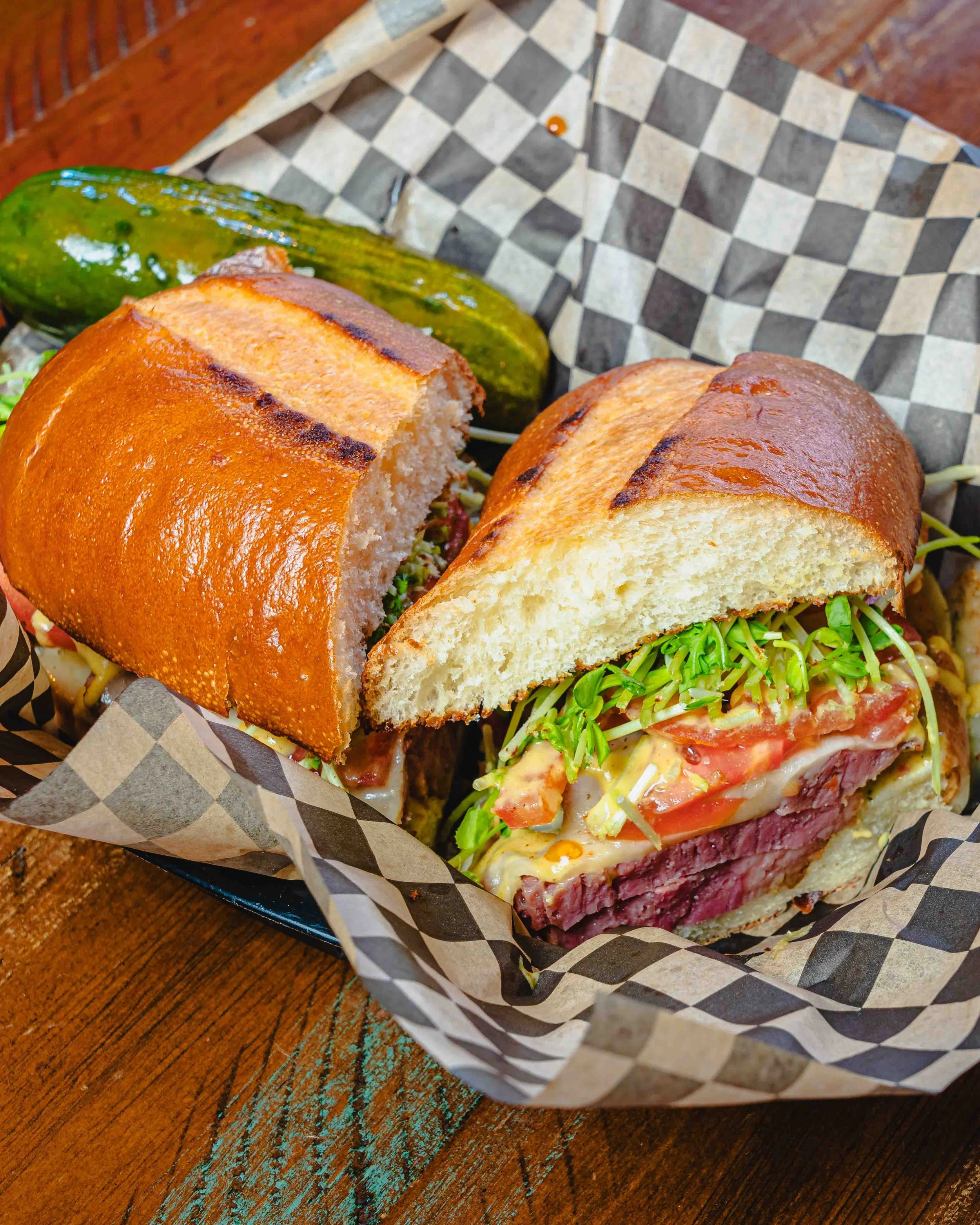 A sandwich with sliced roast beef, tomato, lettuce, and sprouts on a toasted bun, partially cut open, in black and white checkered paper basket on a wooden surface.