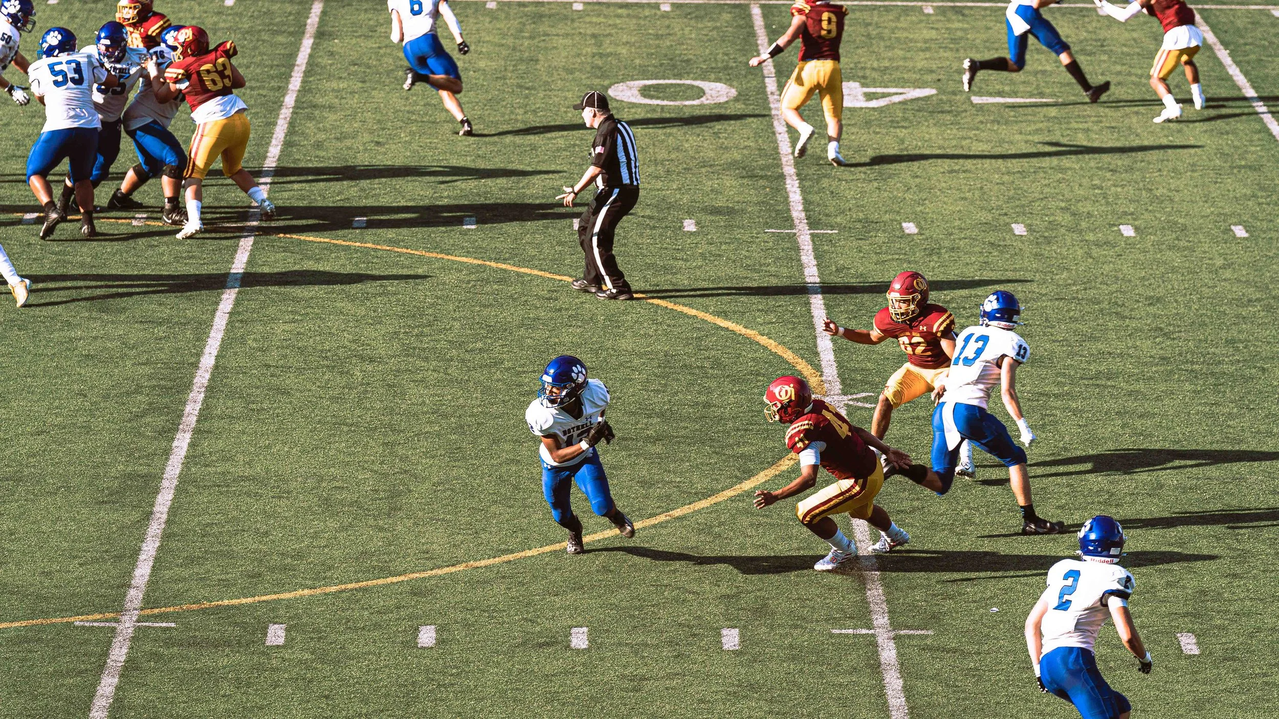 An American football game with players from two teams, one in white and blue uniforms and the other in red and yellow uniforms, on a green field with white yard lines. The players are engaged in a play, with some running and others blocking. An offic