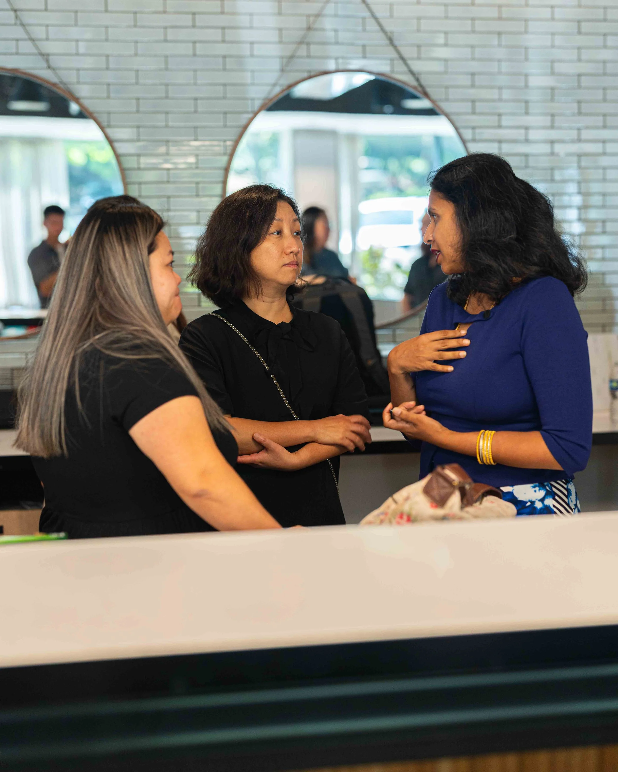 Three women engaged in conversation indoors, with a mirror reflecting other people in the background.