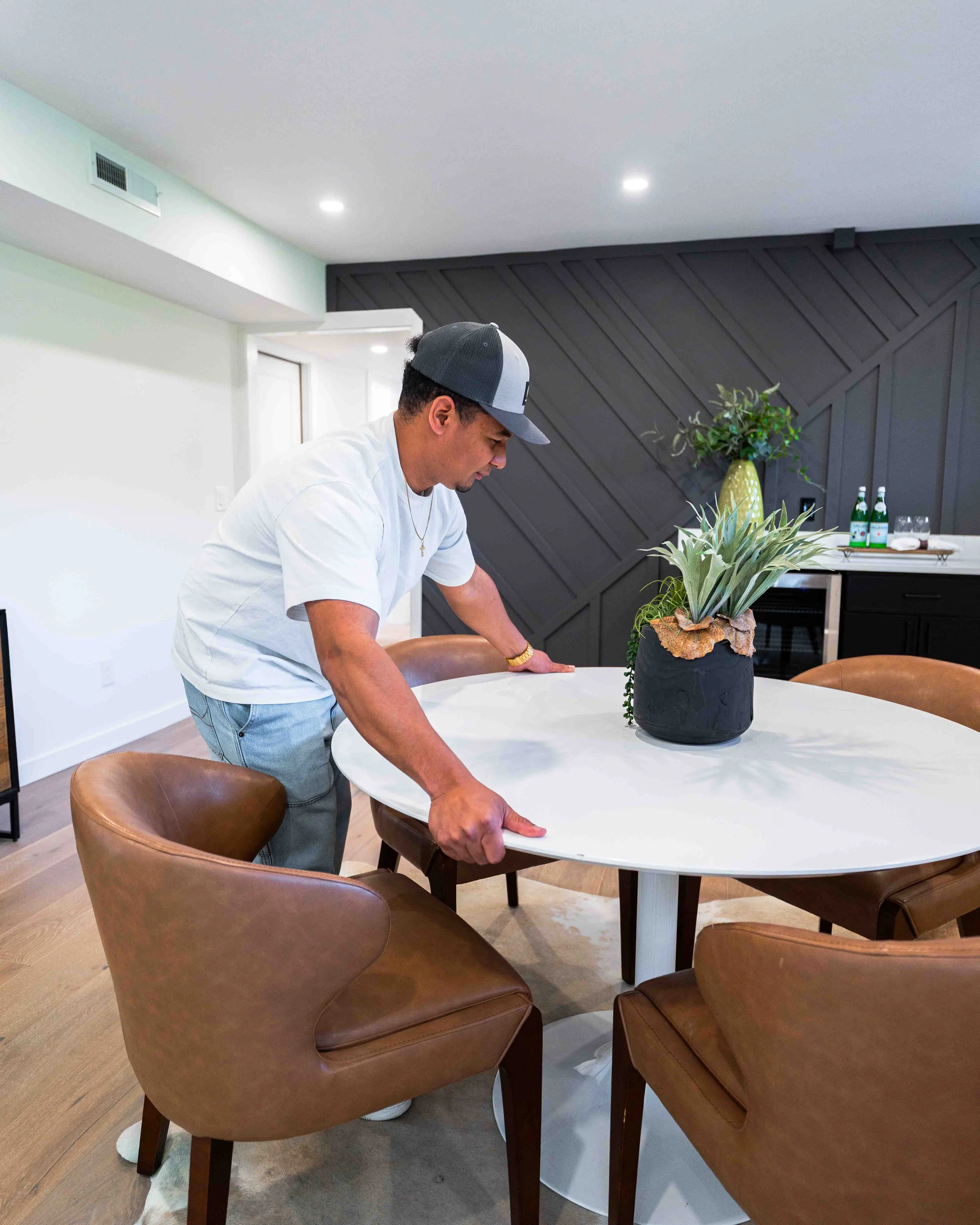 A man in a white shirt and gray cap is leaning over a round white table, adjusting or cleaning it. On the table is a dark-colored pot with a green plant. The background includes a dark gray wall with a geometric pattern, a yellow vase with a green pl