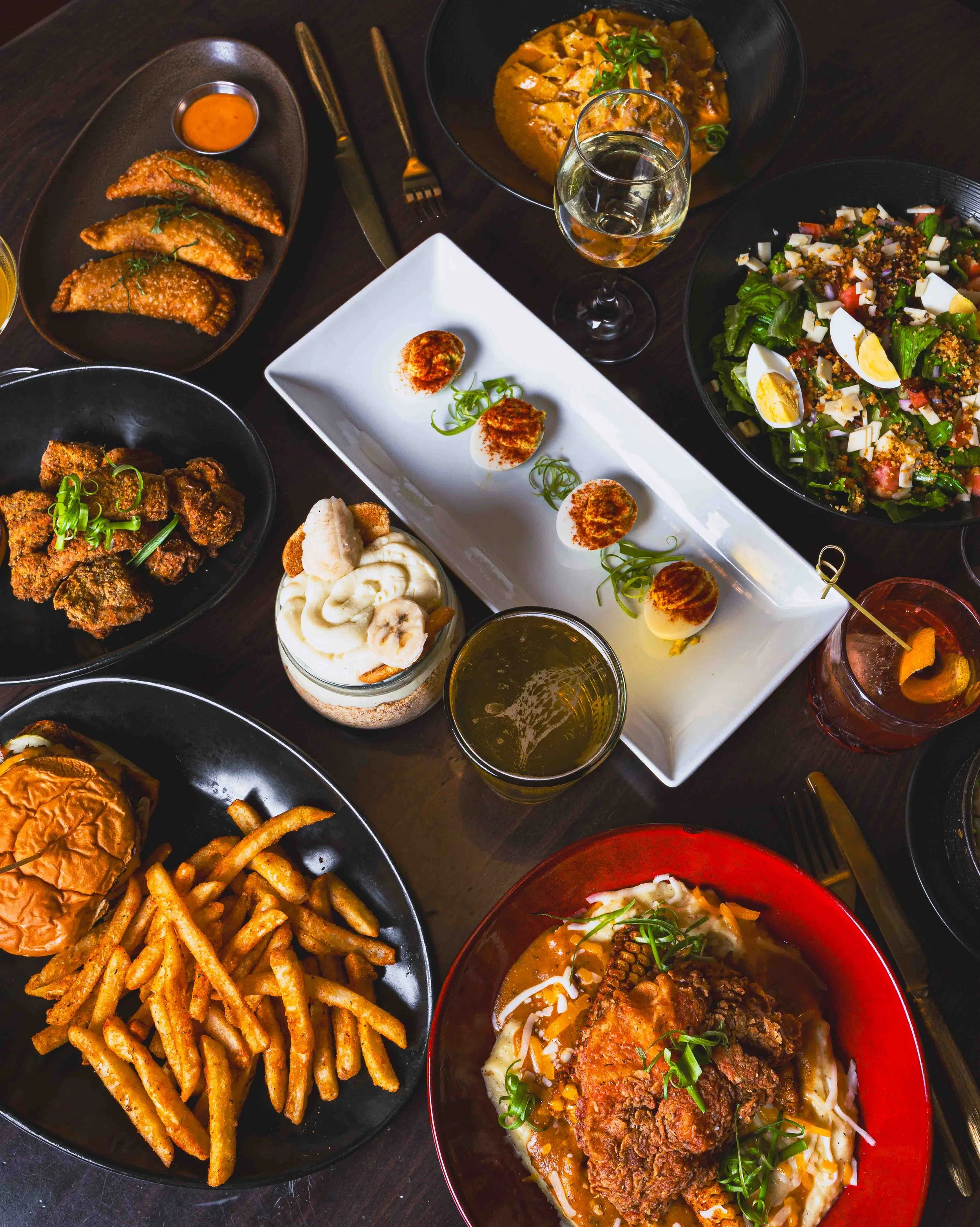 A variety of dishes on a dark table, including French fries, fried chicken, salad with boiled eggs, a bowl of curry, and steamed eggs, with glasses of drinks.