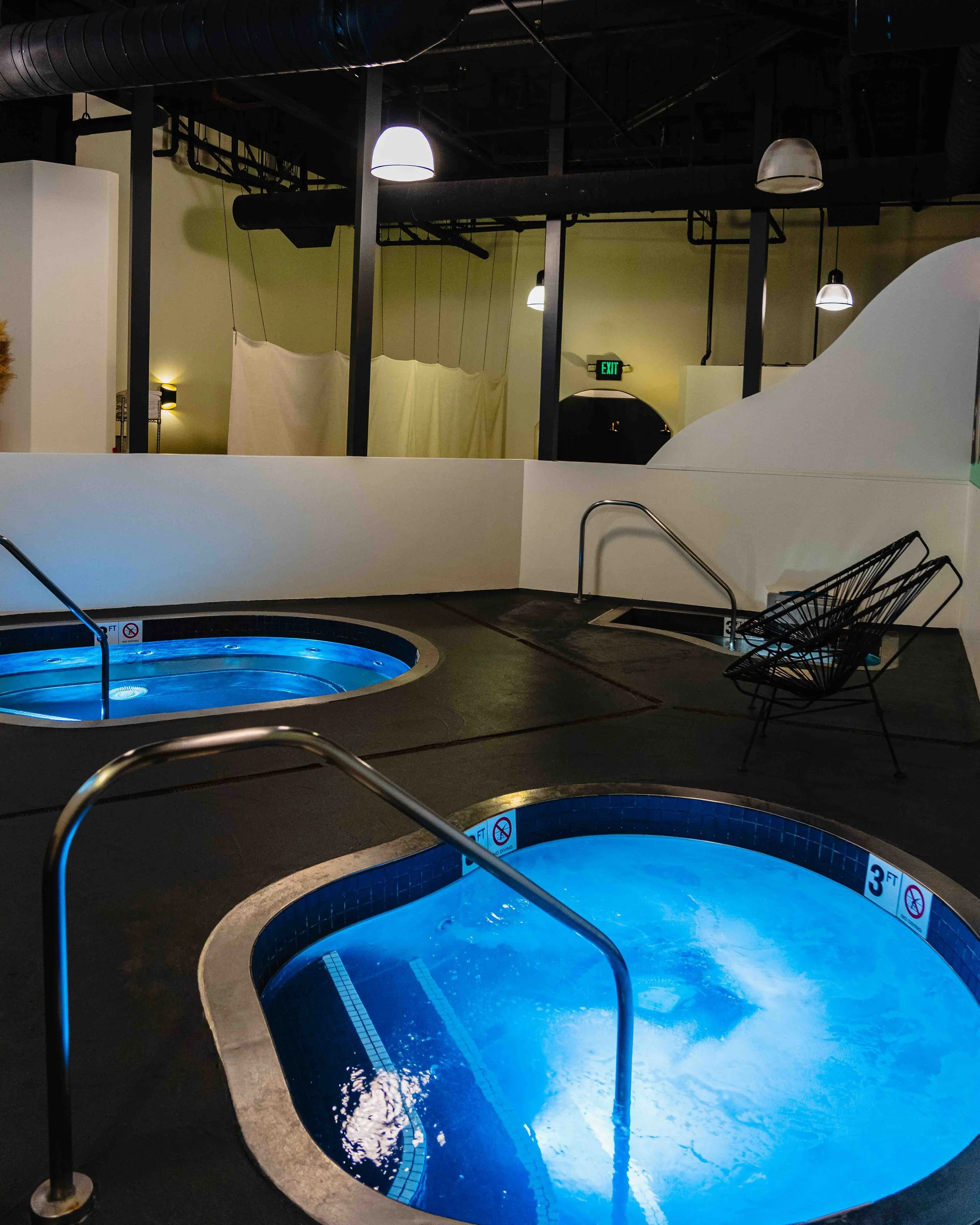 Indoor swimming pool with circular hot tubs filled with blue water, surrounded by black flooring and separated by white barriers. The background shows industrial-style ceiling with black pipes and hanging lights. There are lounge chairs and an exit d