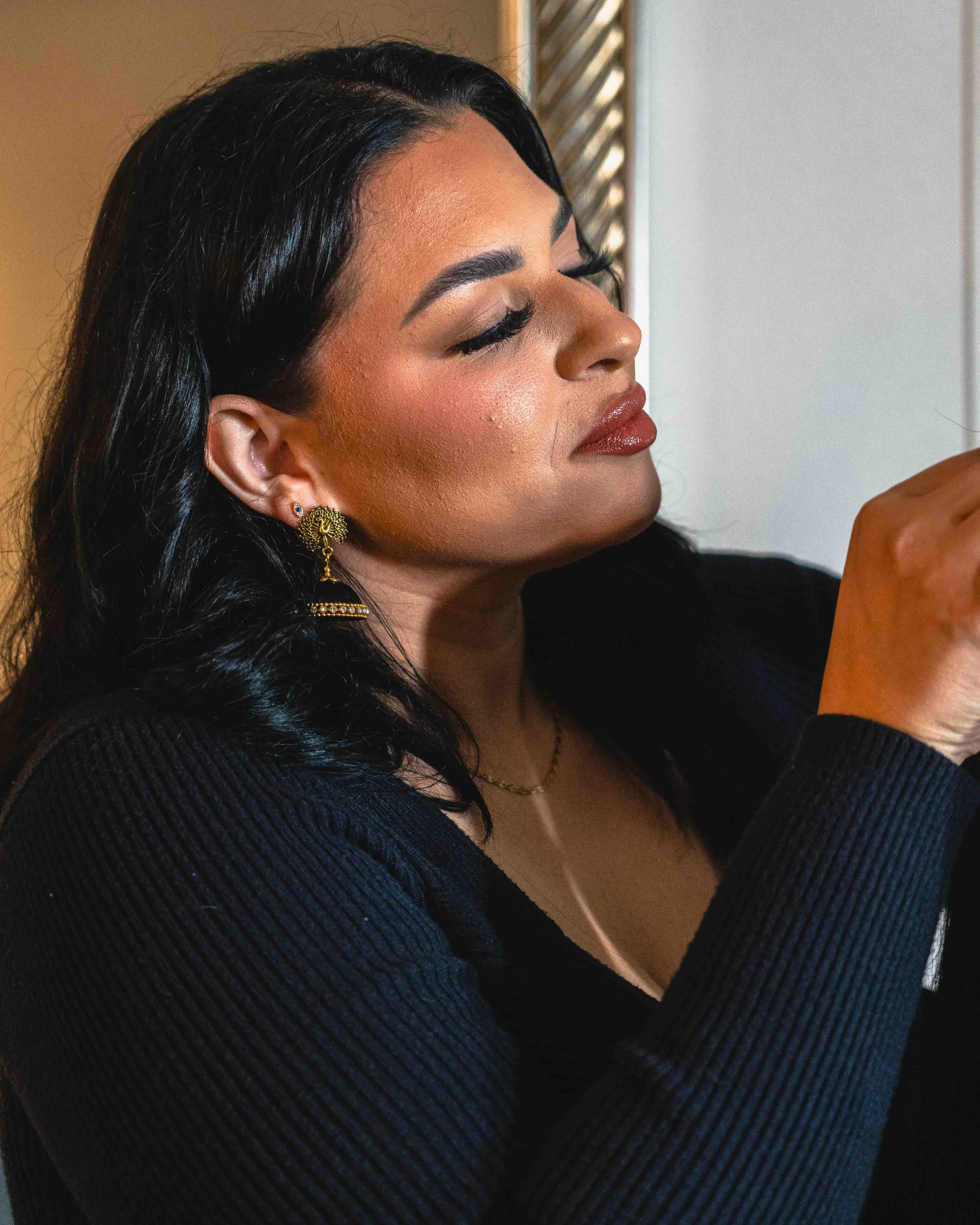 A close-up side profile of a woman with long dark wavy hair, wearing makeup including lipstick, eyeshadow, and earrings, with her eyes closed and hands together in front of her.