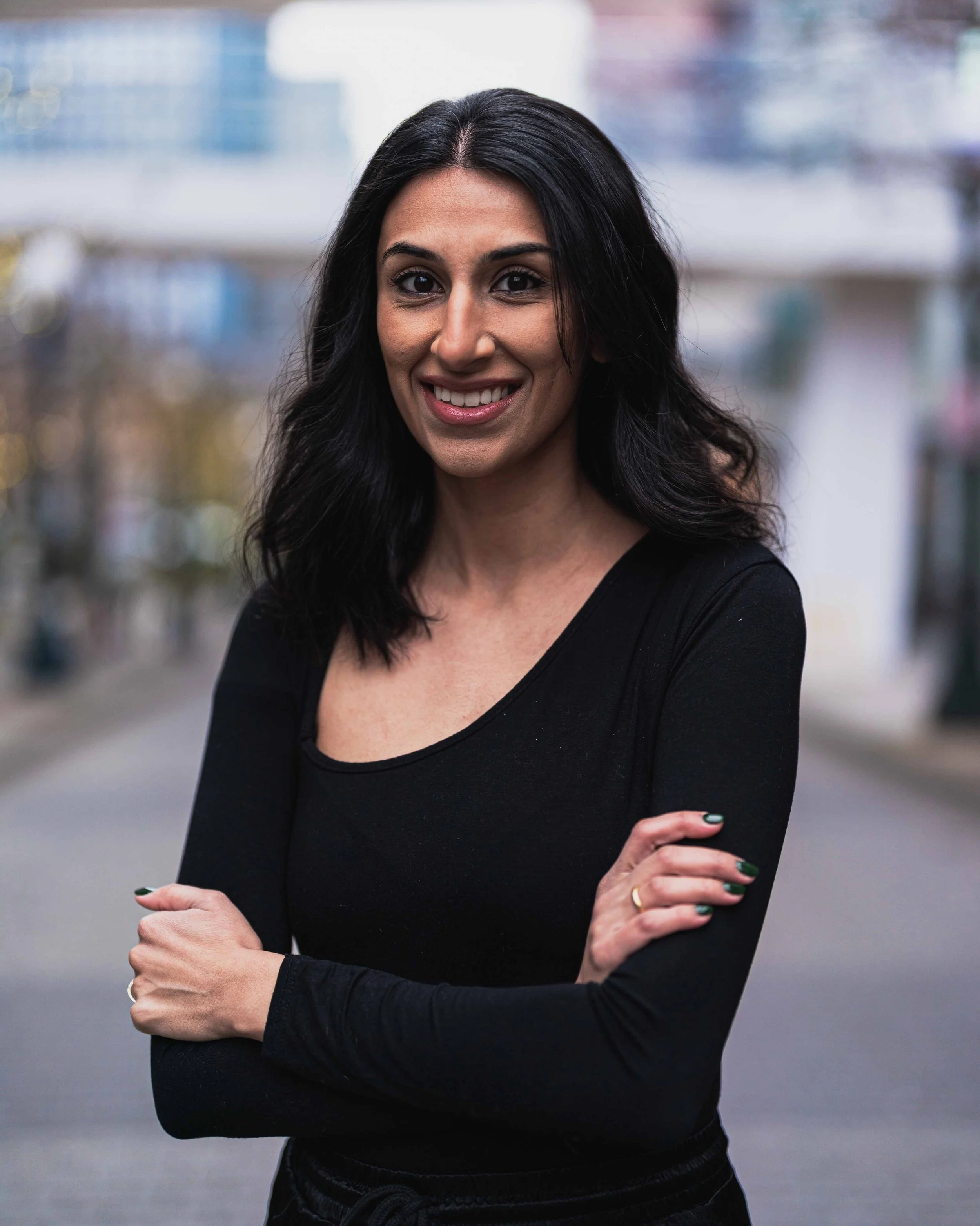 A woman with long black hair, wearing a black top, smiling with arms crossed, in a blurred outdoor setting.