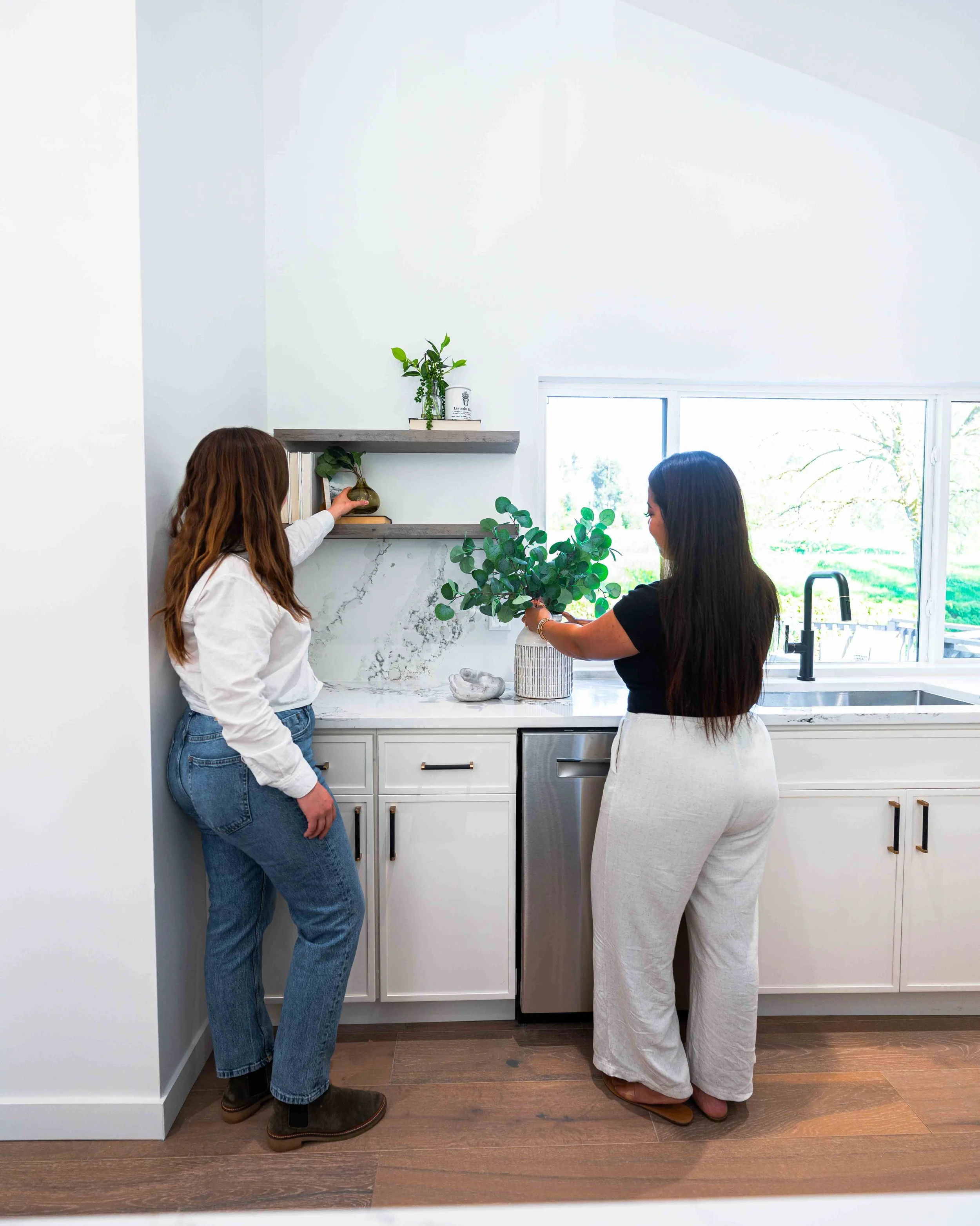 Two women are standing in a kitchen, looking at a potted plant on a countertop. One woman with brown curly hair is in a white shirt and blue jeans, reaching toward the plant. The other woman with straight black hair is in a black shirt and white pant