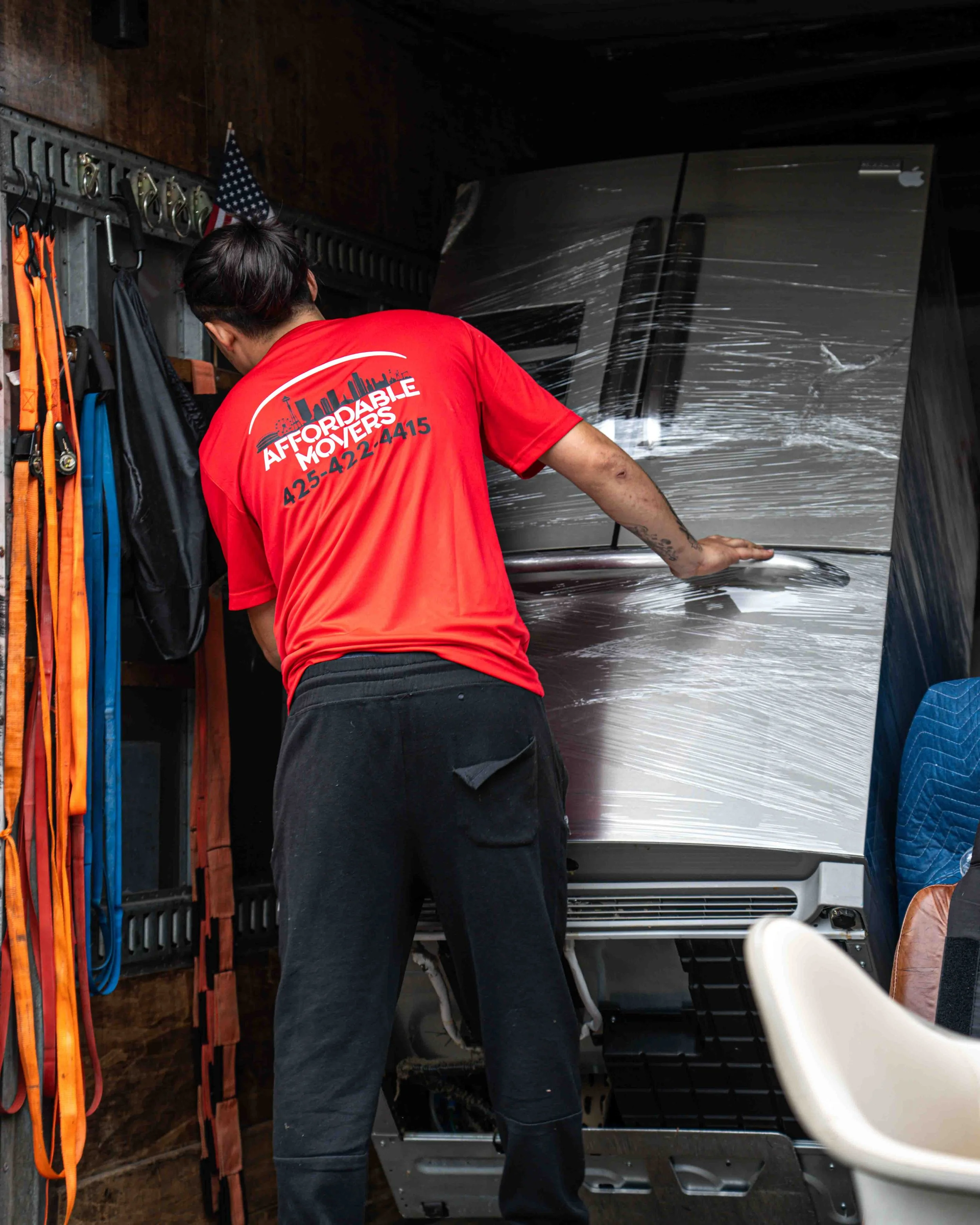 A man wearing a red shirt with the words "Affordable Movers" and a phone number printed on the back is standing in front of a truck, wrapped in plastic, inside a garage or storage area. There are various tools and equipment hanging on the wall to the