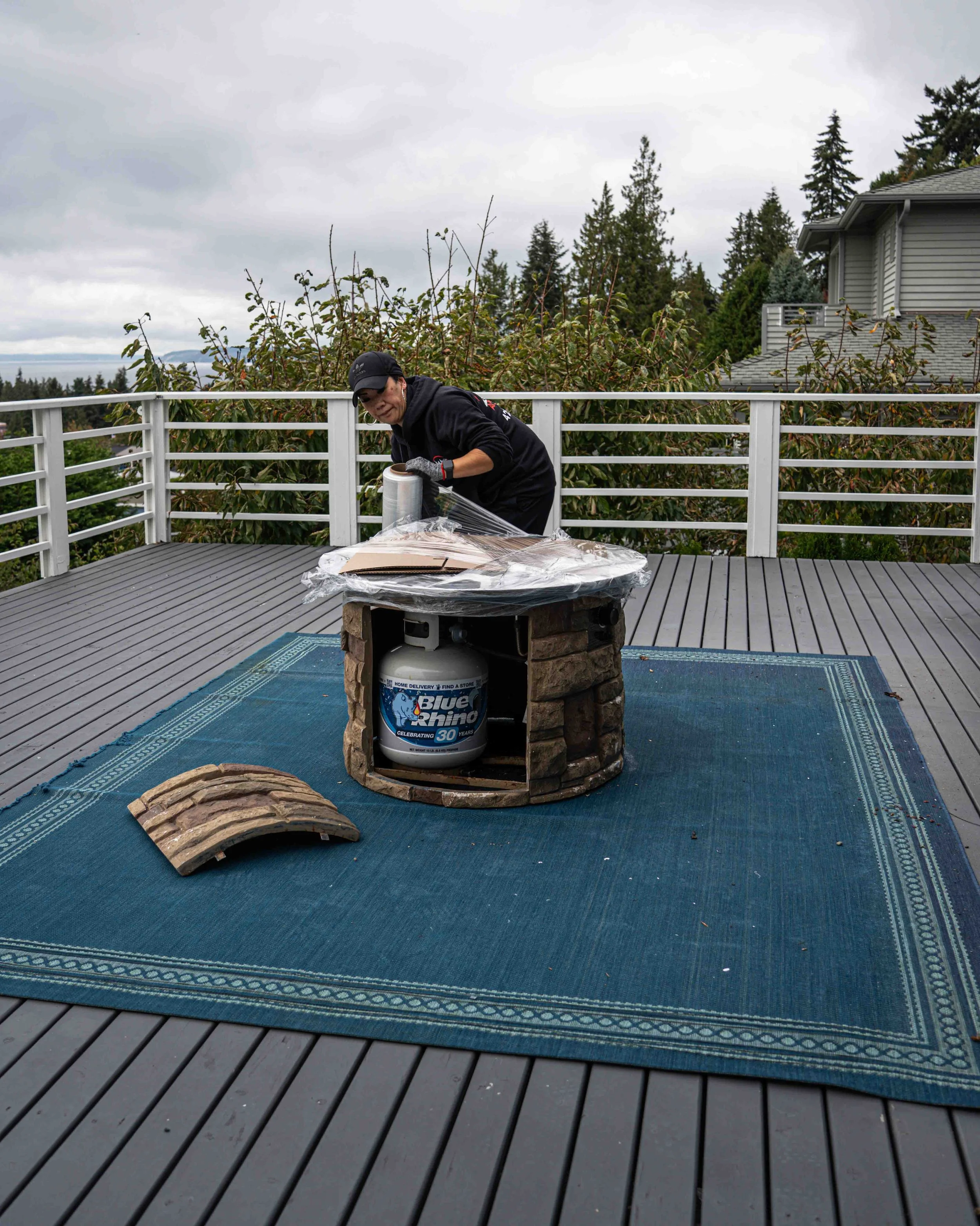 A woman working on a woodworking project on an outdoor deck. She is holding a roll of painter's tape and is standing next to a partly completed piece of furniture. The deck has a blue outdoor rug, and a propane tank is used as support for the project